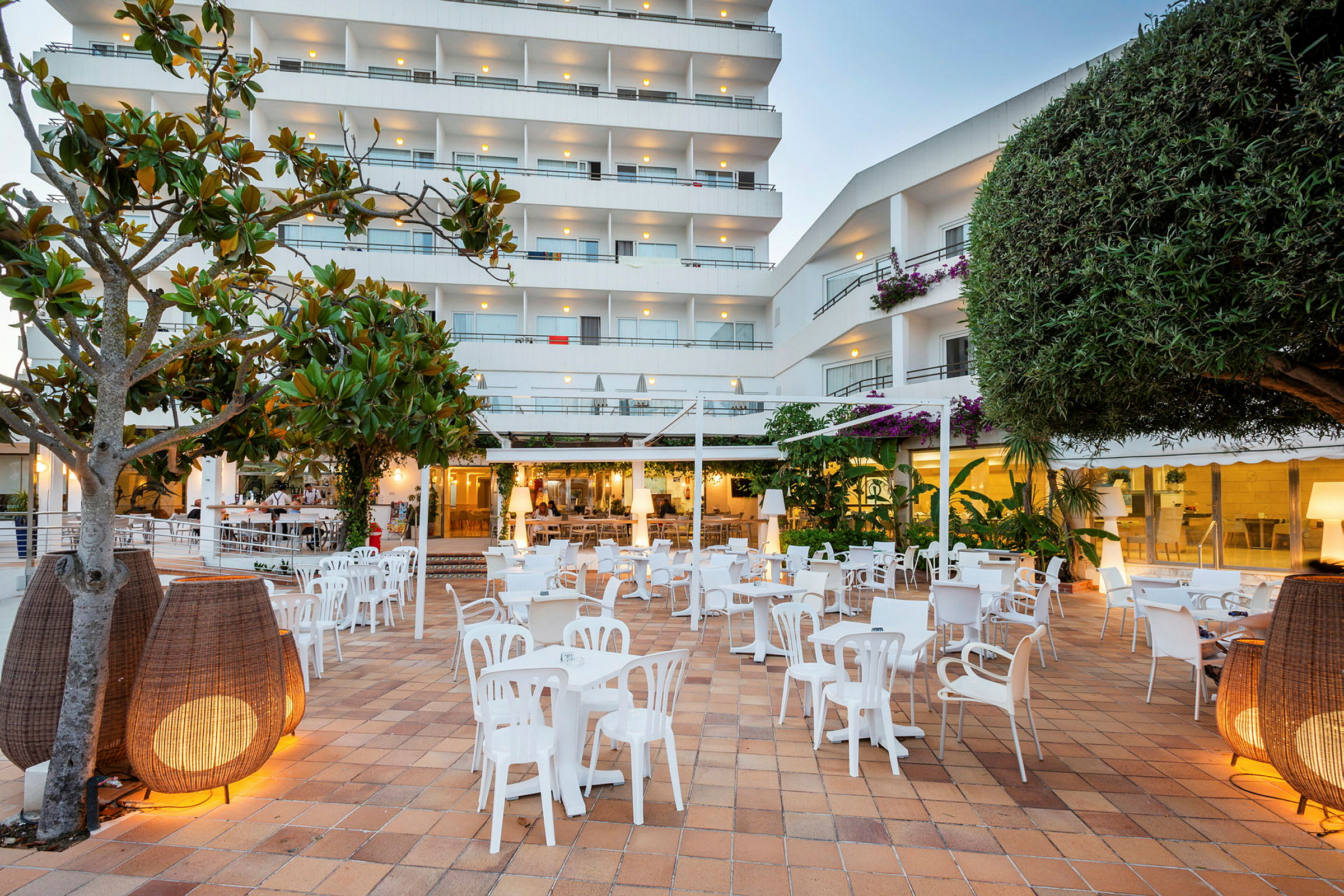 Udendørs område med flere hvide stole og borde, omgivet af planter og moderne belysning, med et flerhushotel i baggrunden. (AI-genereret tekst)