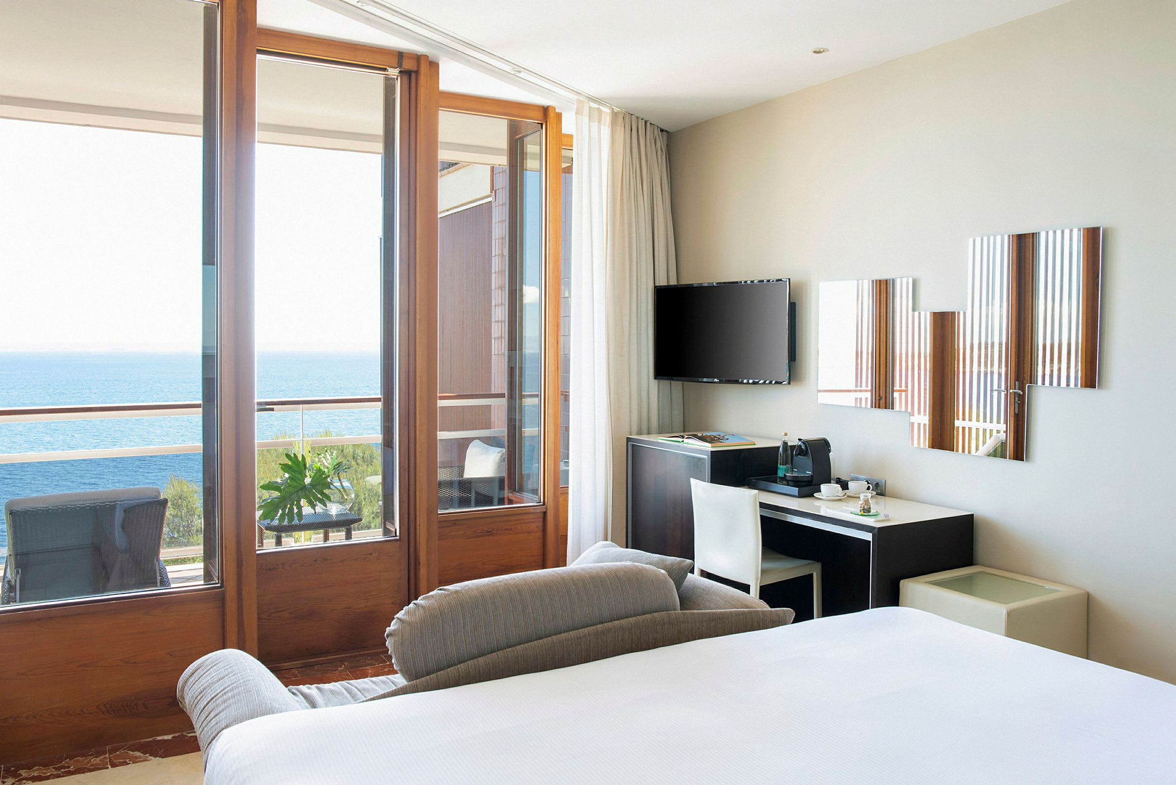 Et moderne hotelværelse med en seng, skrivebord, TV og store glasdøre, der fører ud til en balkon med udsigt over havet. (AI-genereret tekst)