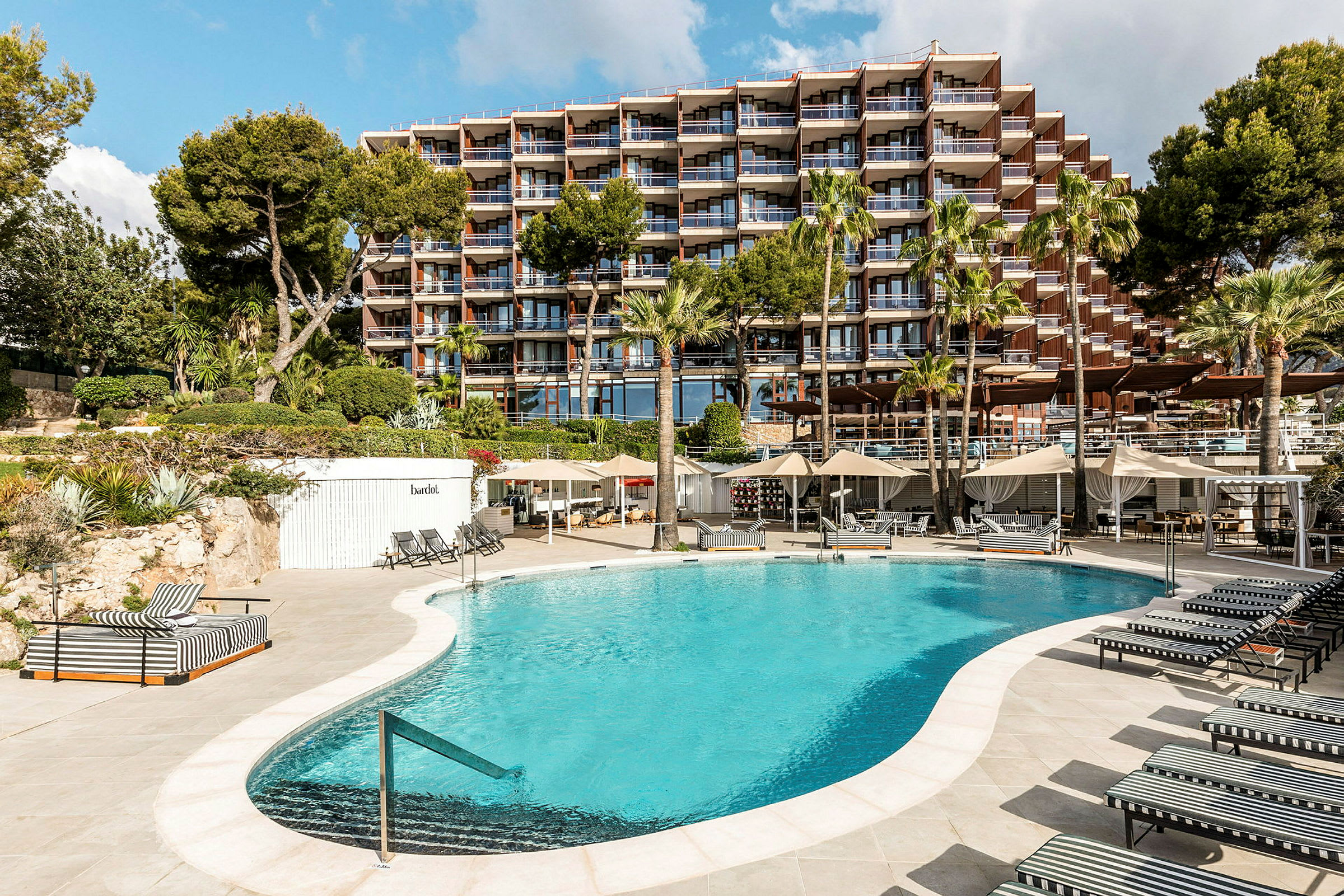 En udendørs swimmingpool omgivet af solsenge og parasoller foran en større hotelbygning. (AI-genereret tekst)