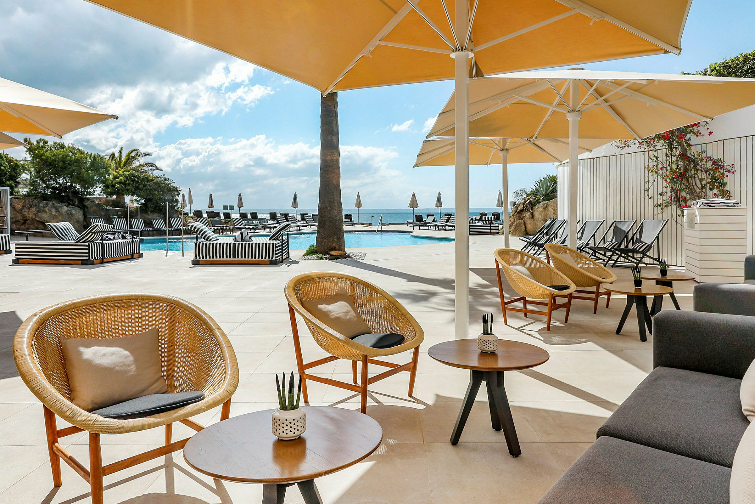 Billede af en poolterrasse med parasoller, stole og sofaer. En klar blå himmel og træer i baggrunden. (AI-genereret tekst)