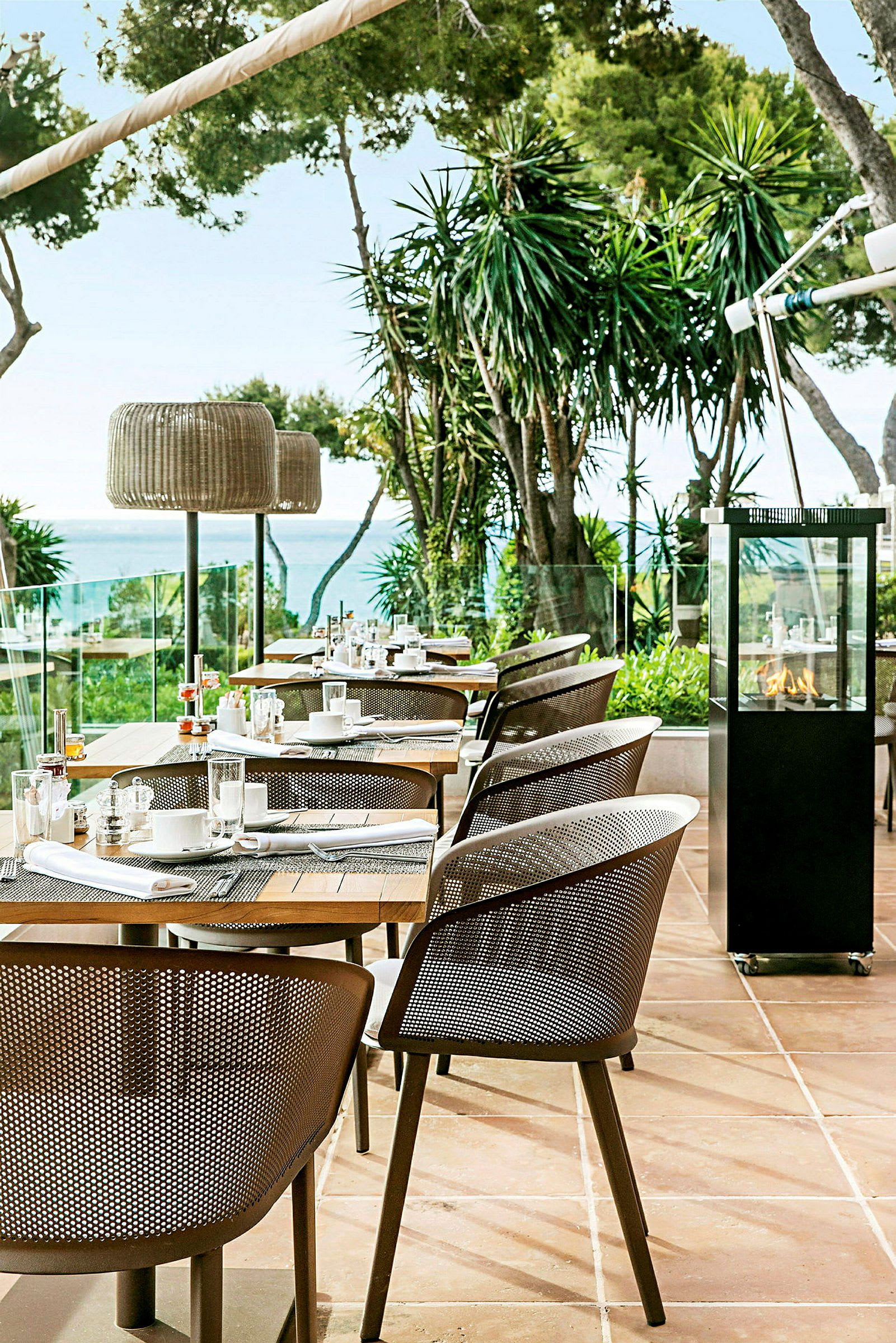 Udendørsrestaurant med moderne stole og dækkede borde, omgivet af grønne områder og udsigt til havet. (AI-genereret tekst)
