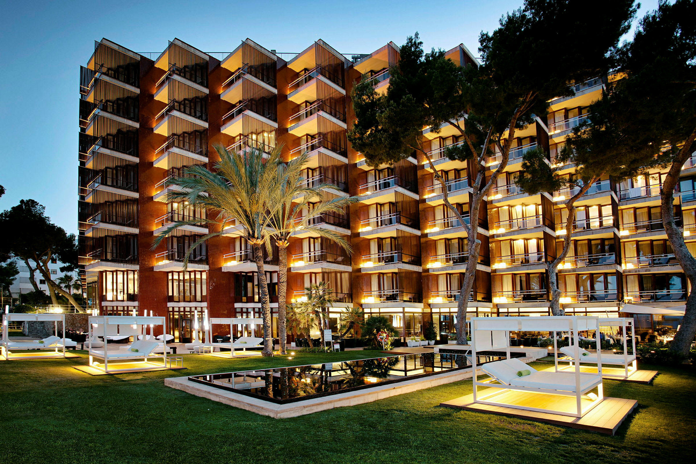 Et hotel i skumringen med oplyste altaner og en frodig have med palmer og moderne solbænke. (AI-genereret tekst)