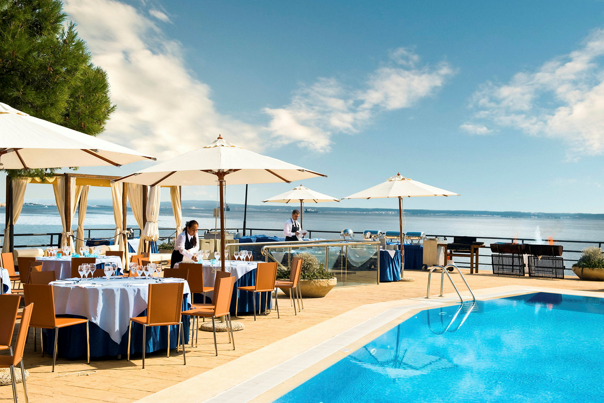 Restaurantområde ved en pool med udsigt over havet. Bord og parasoller er opsat. (AI-genereret tekst)