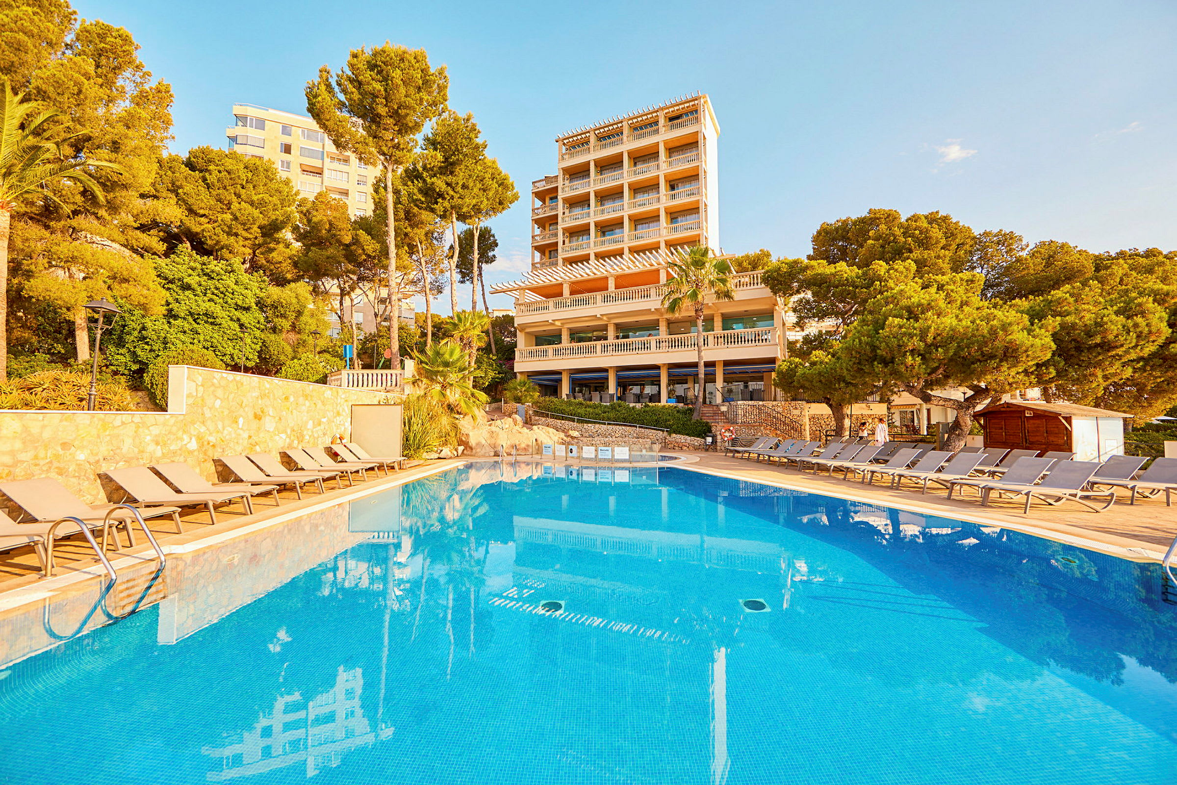 Udendørs pool foran hotelbygning, omgivet af liggestole og grønt. (AI-genereret tekst)