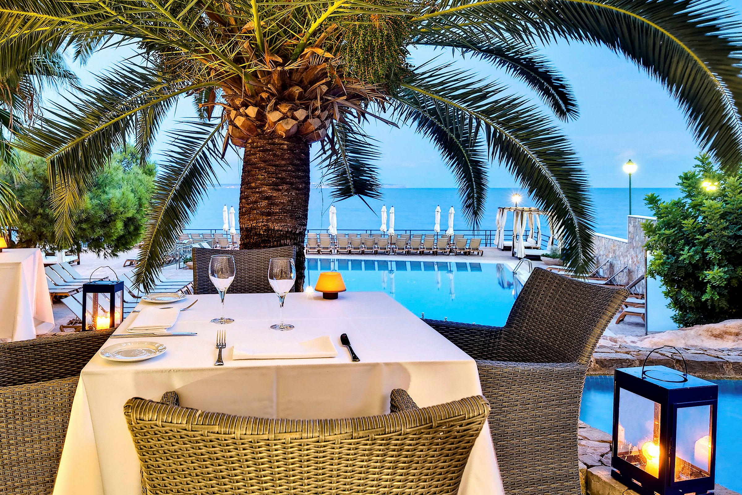 Udsigt over en restaurant ved en pool nær havet med en palme i forgrunden. (AI-genereret tekst)