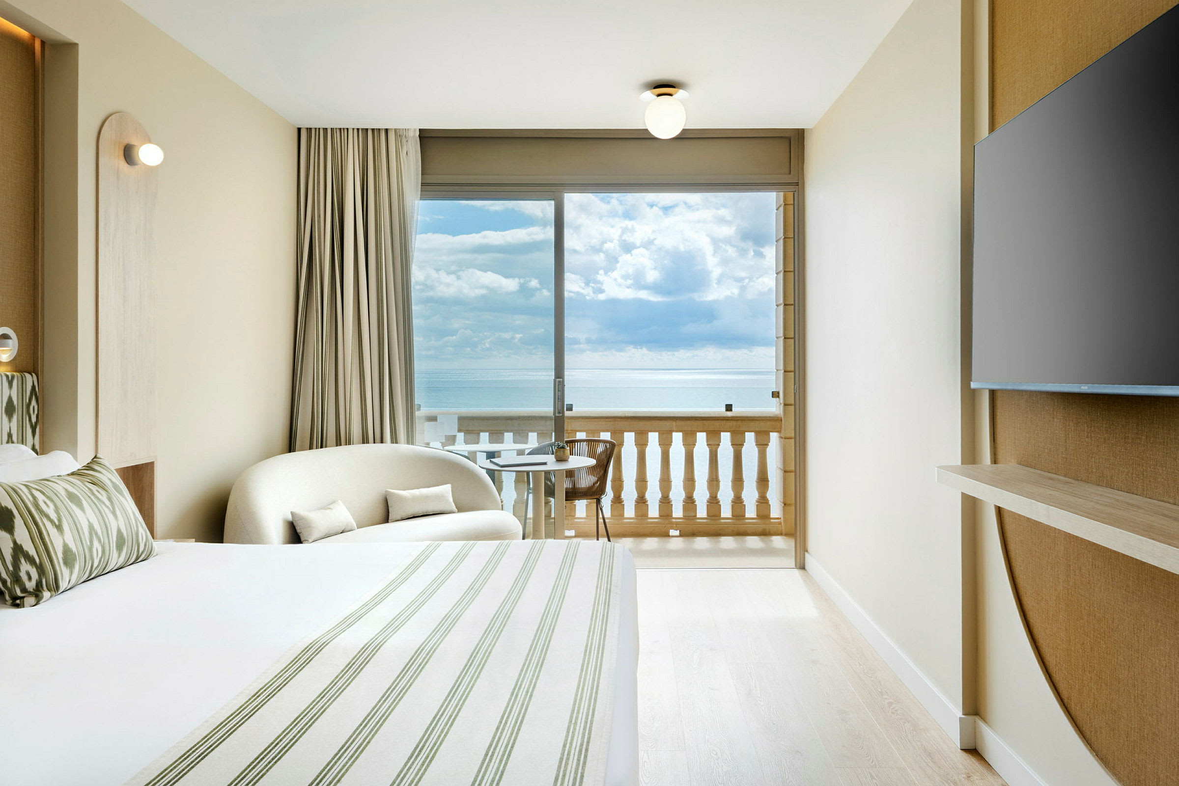 Et lyst hotelværelse med en seng, en siddeplads, et TV på væggen og en balkon med udsigt over havet. (AI-genereret tekst)
