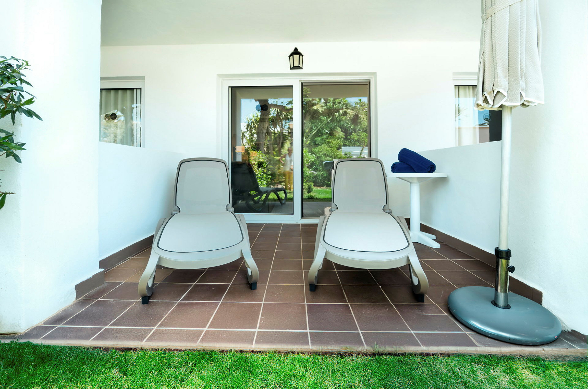 To liggestole på en terrasse ved et hotelværelse, med et bord og en sammenfoldet parasol. (AI-genereret tekst)