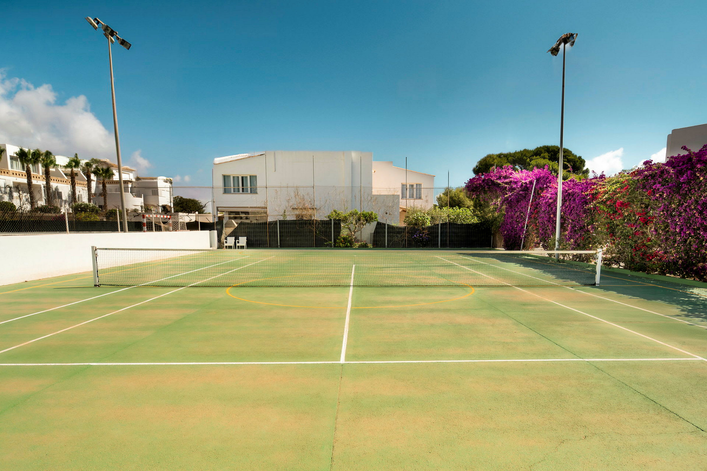 En tennisbane med grøn overflade omgivet af bygninger og beplantning. (AI-genereret tekst)