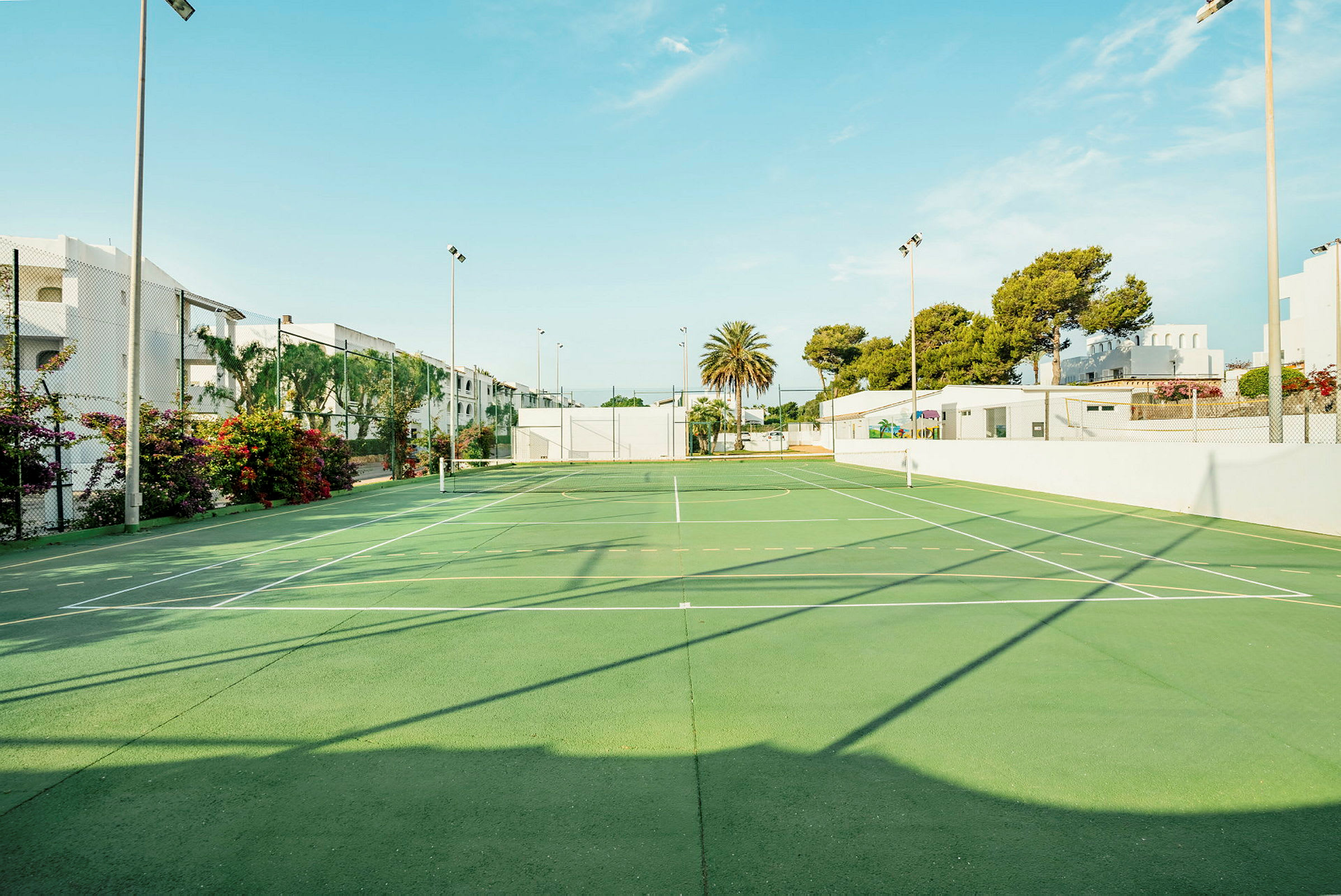 En grøn tennisbane udendørs med net, omgivet af hvide bygninger, træer og blomster under en klar himmel. (AI-genereret tekst)