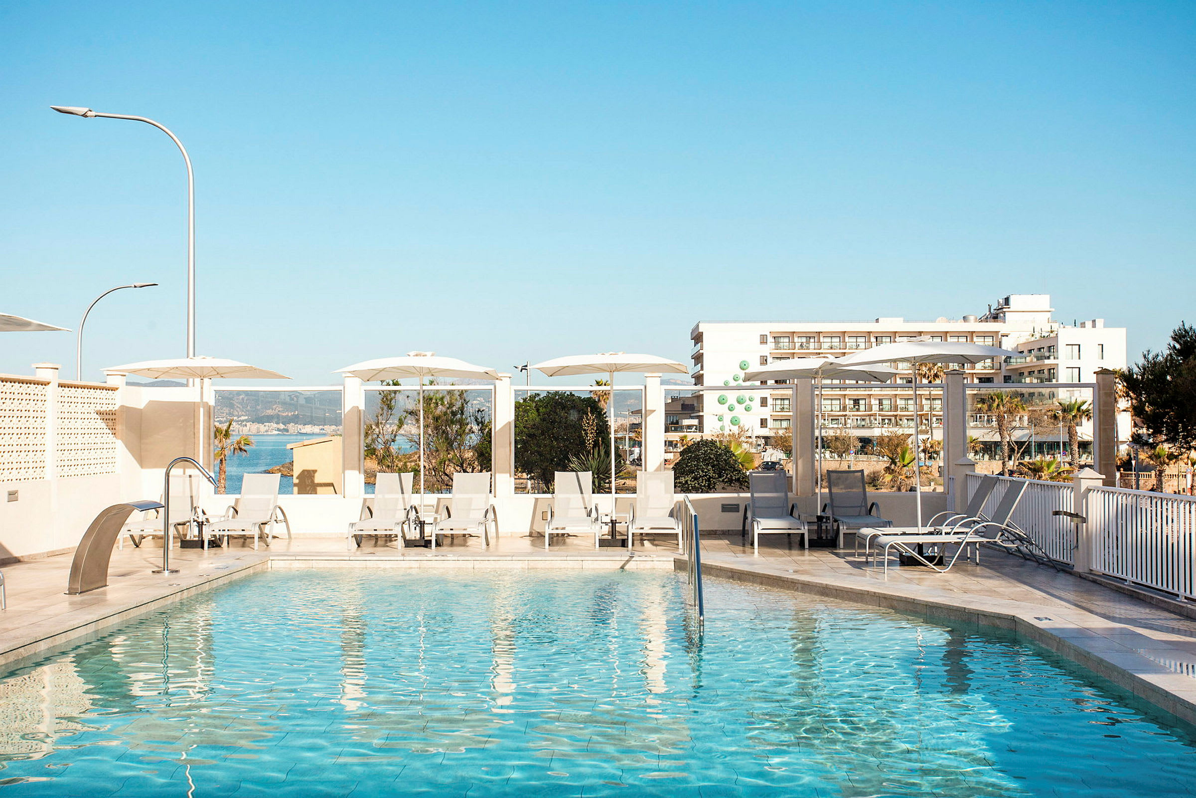 Udendørs pool med liggestole og parasoller, i baggrunden ses bygninger og havet. (AI-genereret tekst)