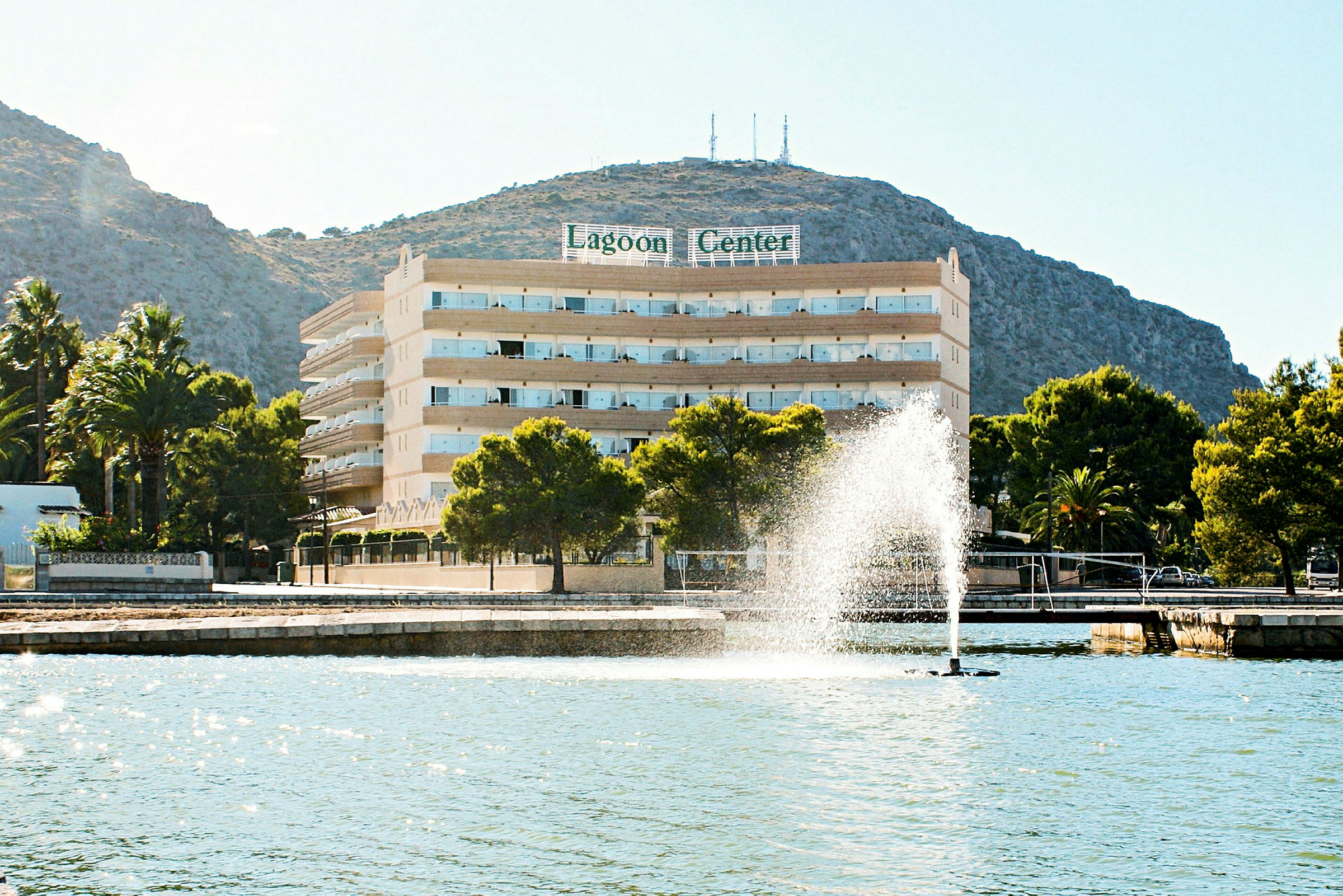 Hotel Lagoon Center ved strandpromenaden med en fontæne og bjerge i baggrunden. (AI-genereret tekst)