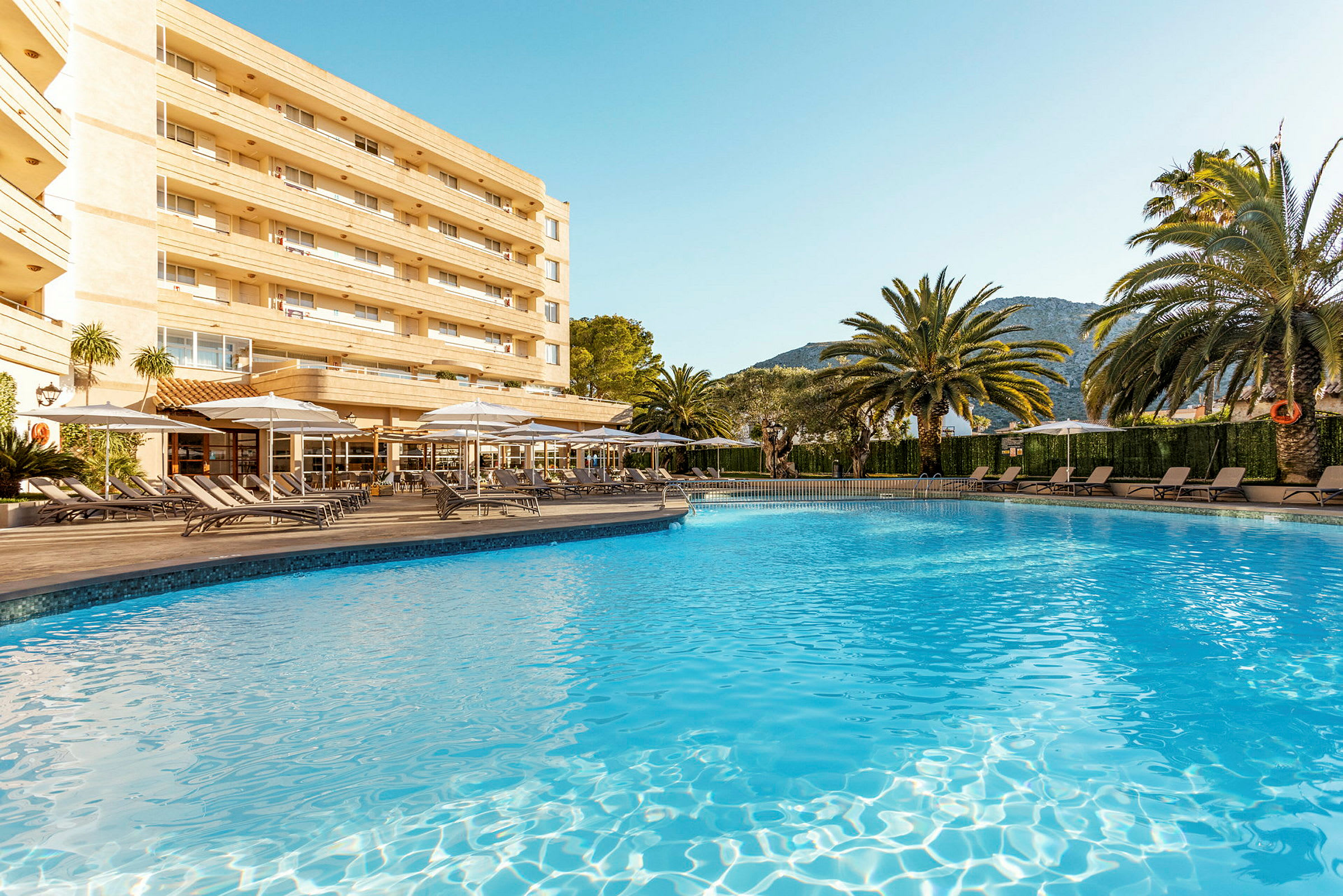Billede af en pool med liggestole og et hotel i baggrunden. (AI-genereret tekst)