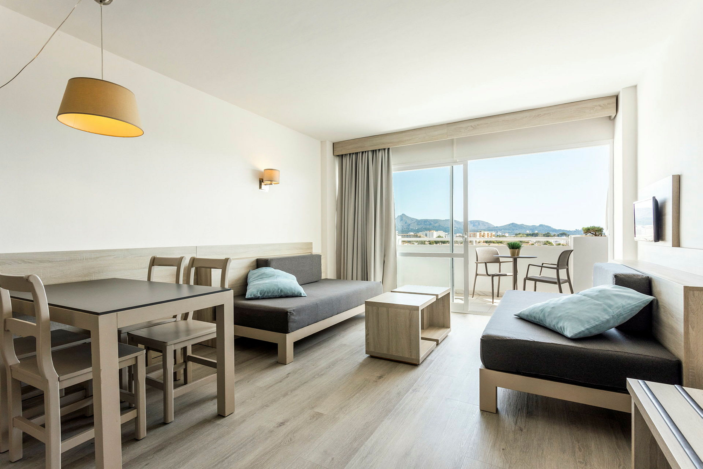 Et lyst hotelværelse med spisebord og stole, sofaer samt en balkon med udsigt til bjergene. (AI-genereret tekst)