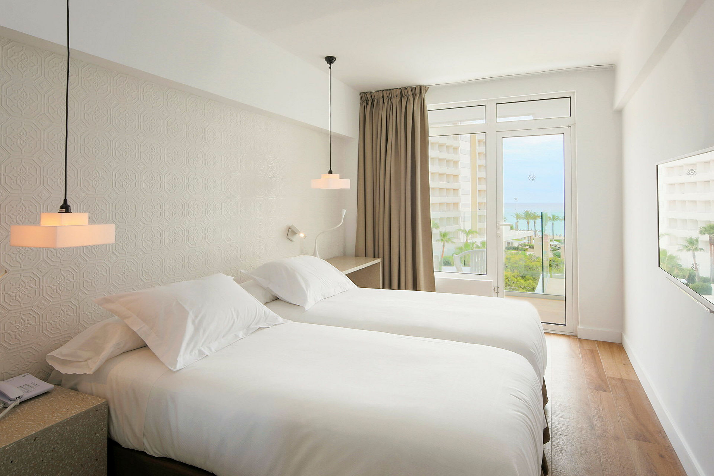 Et hotelværelse med to enkeltsenge, lyst interiør og en balkon med udsigt til havet. (AI-genereret tekst)