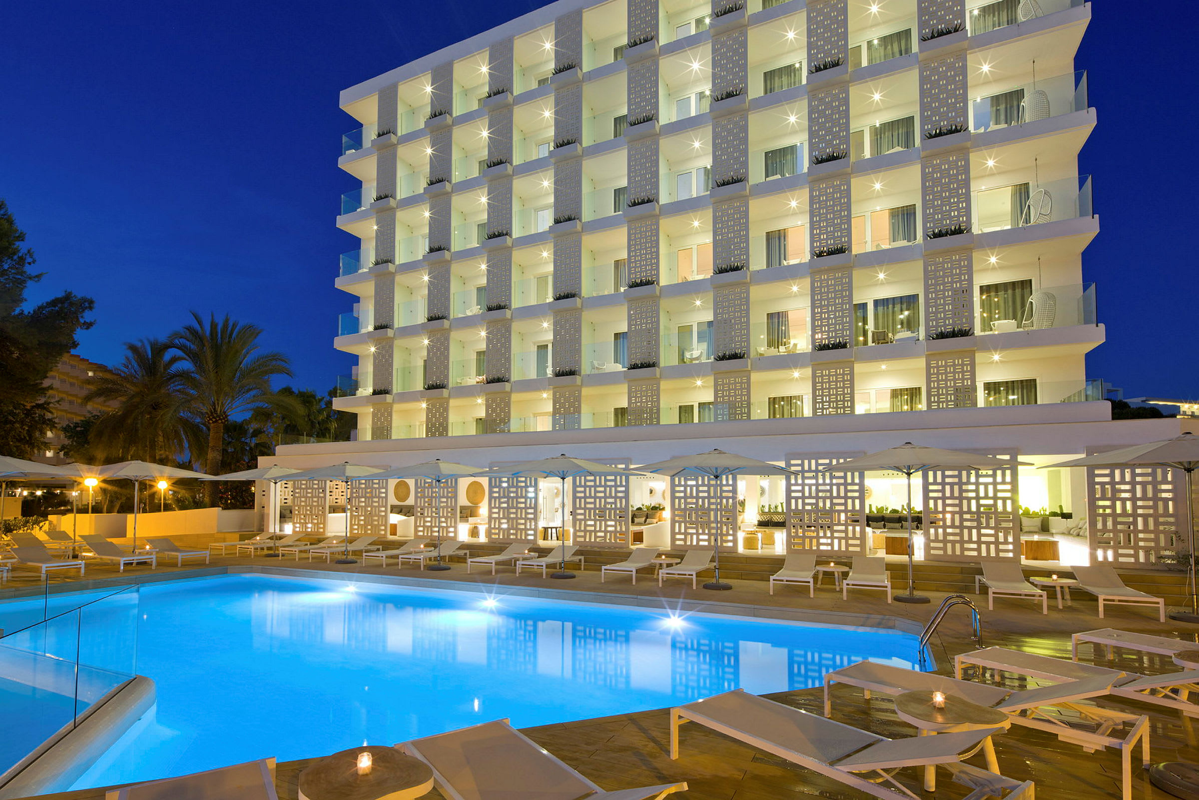 Billede af et hotel med en oplyst pool, liggestole og en moderne facade om aftenen. (AI-genereret tekst)