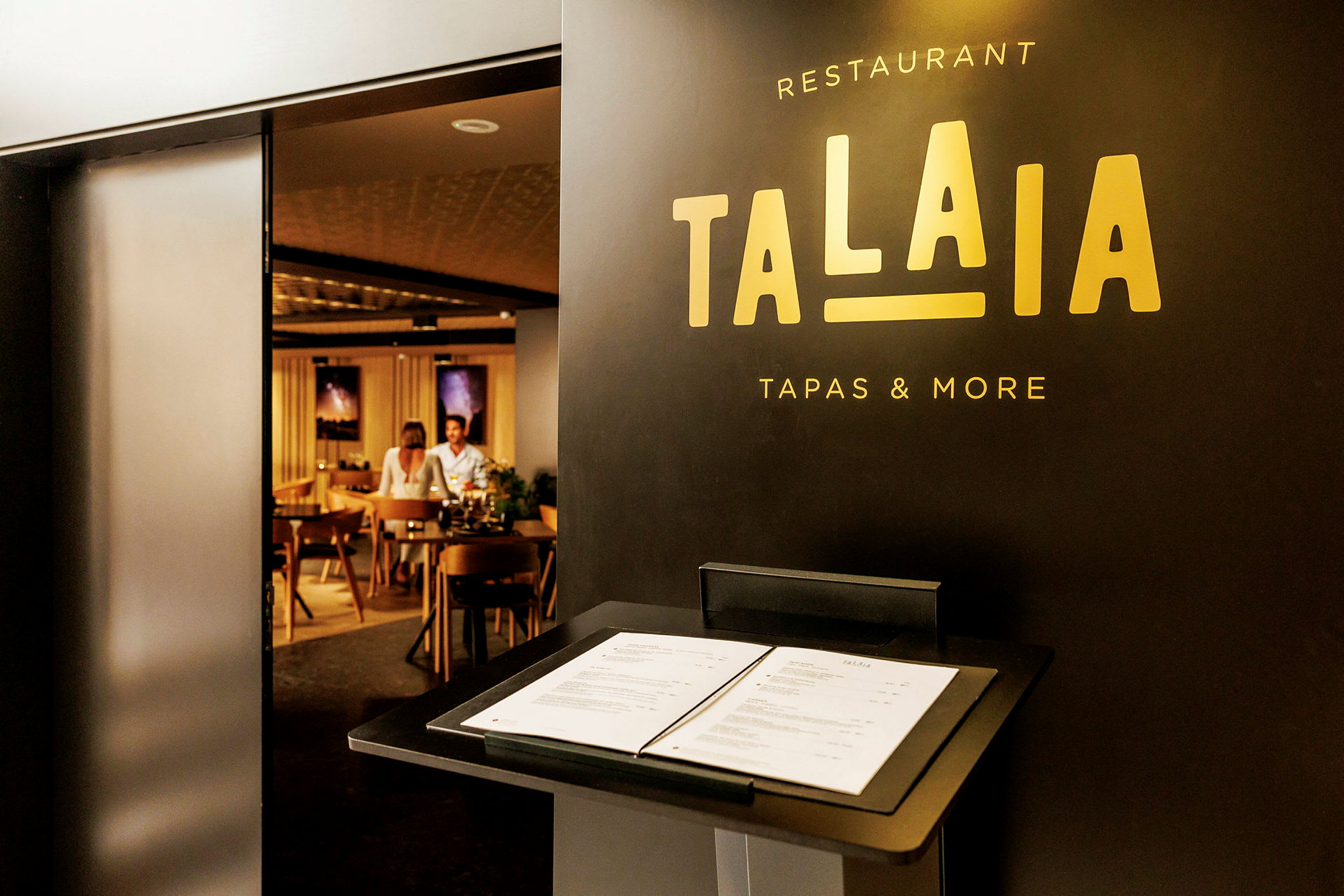 Restaurant Talaia med en menu på et stativ nær indgangen, der viser et kig ind i en stilfuld indretning med siddepladser. (AI-genereret tekst)