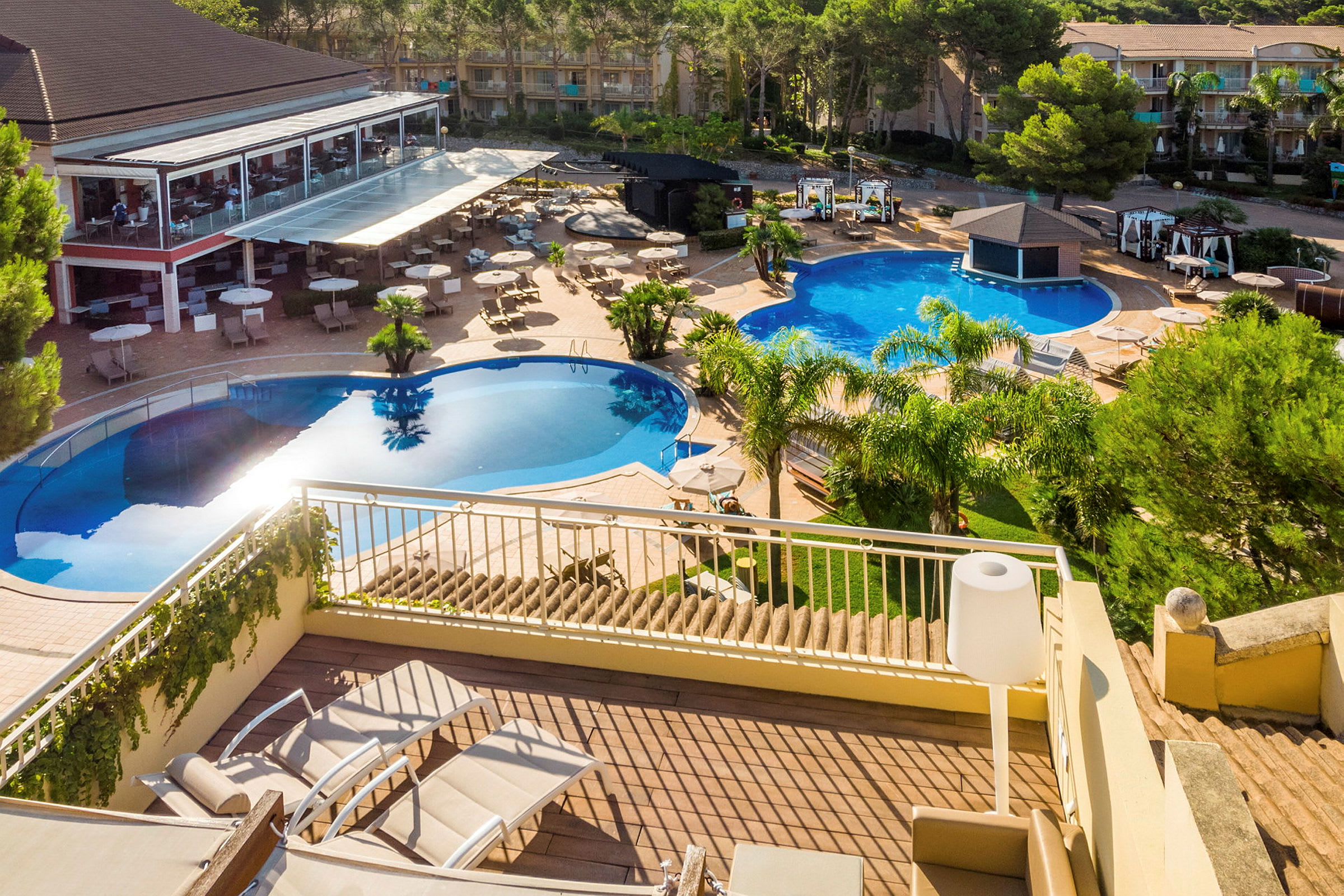 Billede af et udendørsområde med to swimmingpools omgivet af planter og liggestole samt en restaurant med terrasse. (AI-genereret tekst)