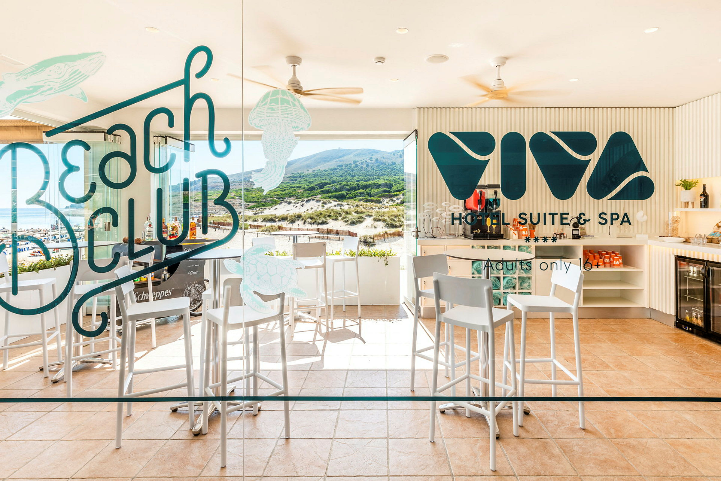 Billede af en hotelbar med høje stole, borde og glasvæg med teksten 'Beach Club' og 'Viva Hotel Suite & Spa' samt udsigt mod en strand og grønne højder. (AI-genereret tekst)