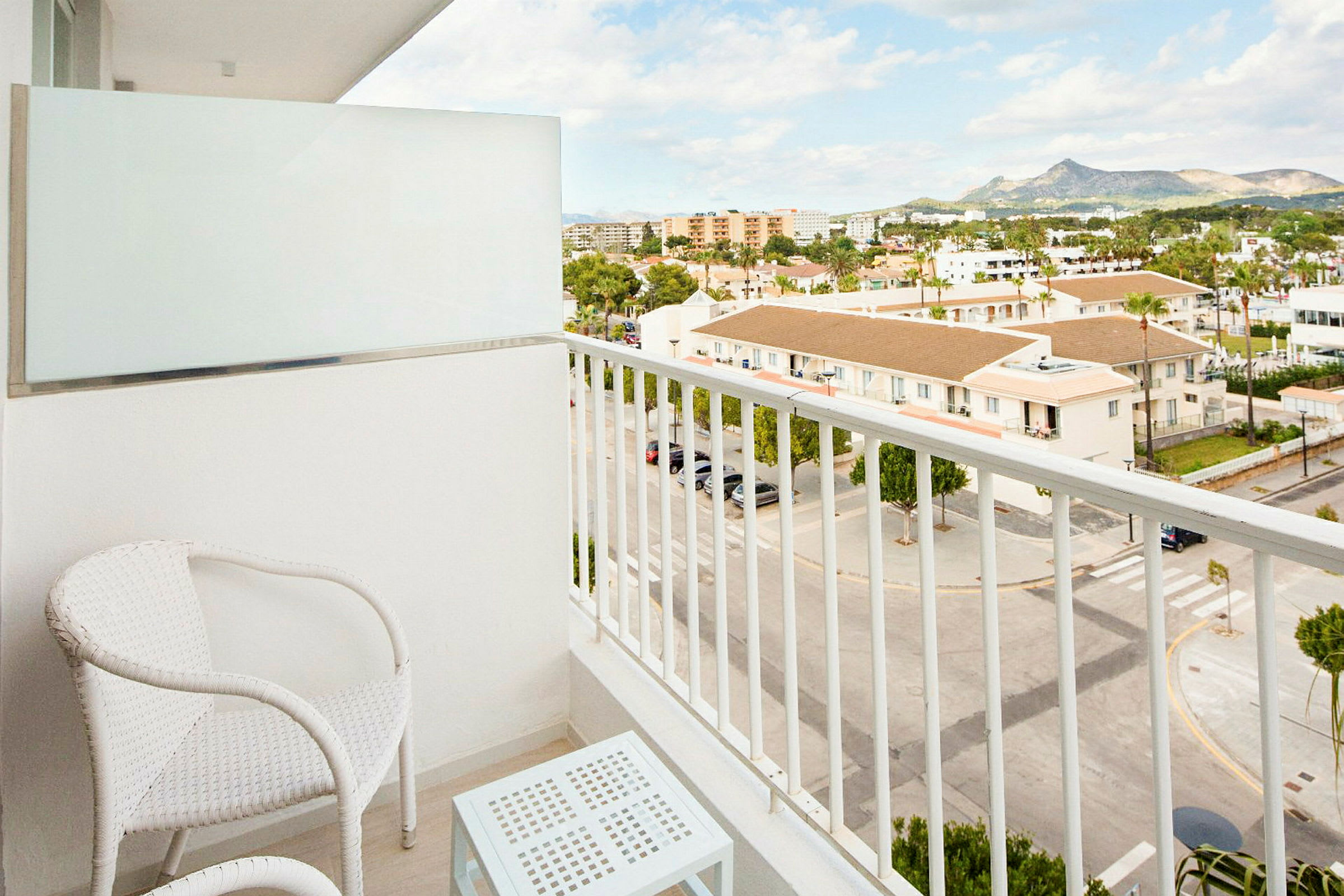 En balkon med to hvide stole og et bord, med udsigt over en gade og bygninger. (AI-genereret tekst)