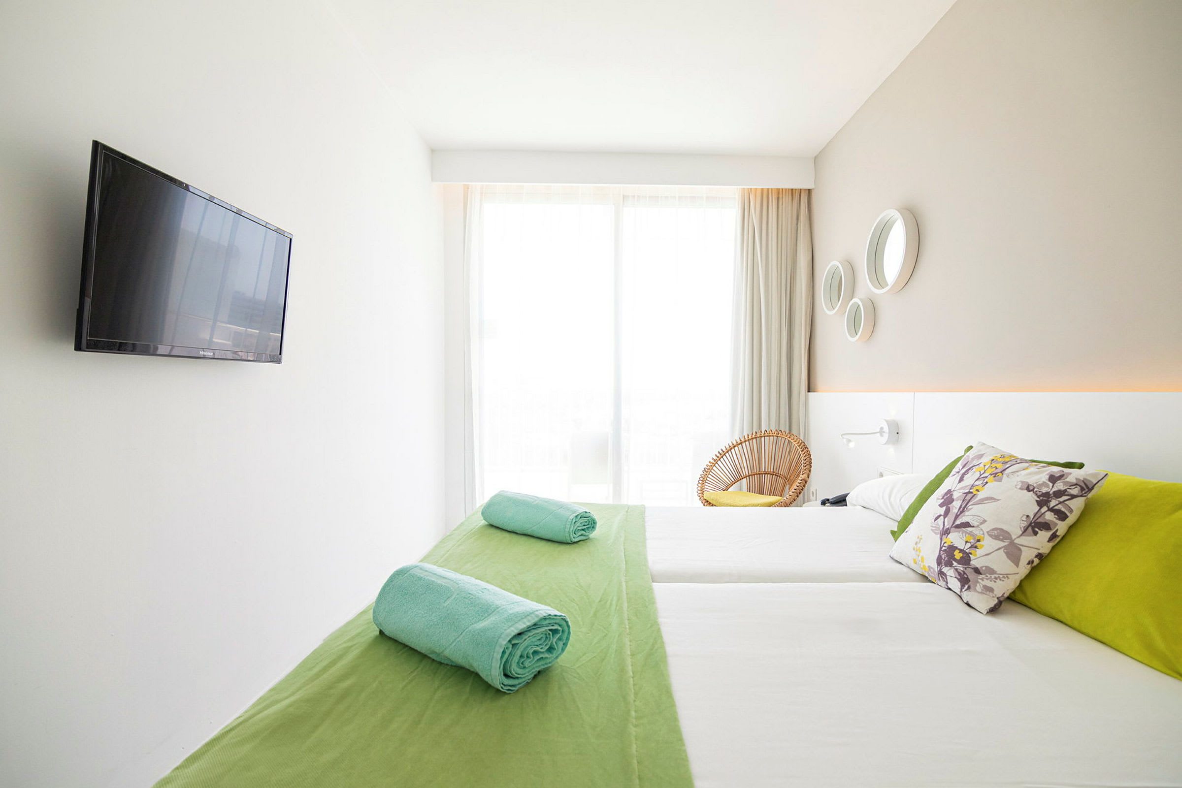 Et lyst hotelværelse med en dobbeltseng, farverige puder, sammenrullede håndklæder og et fladskærms-tv på væggen. (AI-genereret tekst)
