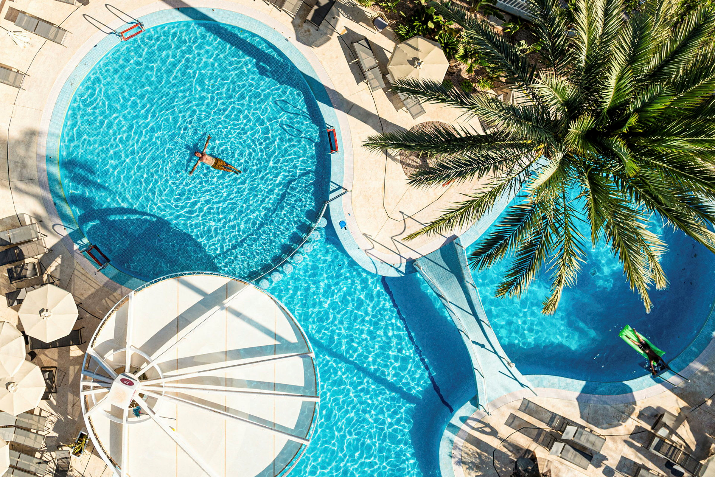 En udendørs pool omgivet af solsenge og parasoller, med en person der svømmer. (AI-genereret tekst)
