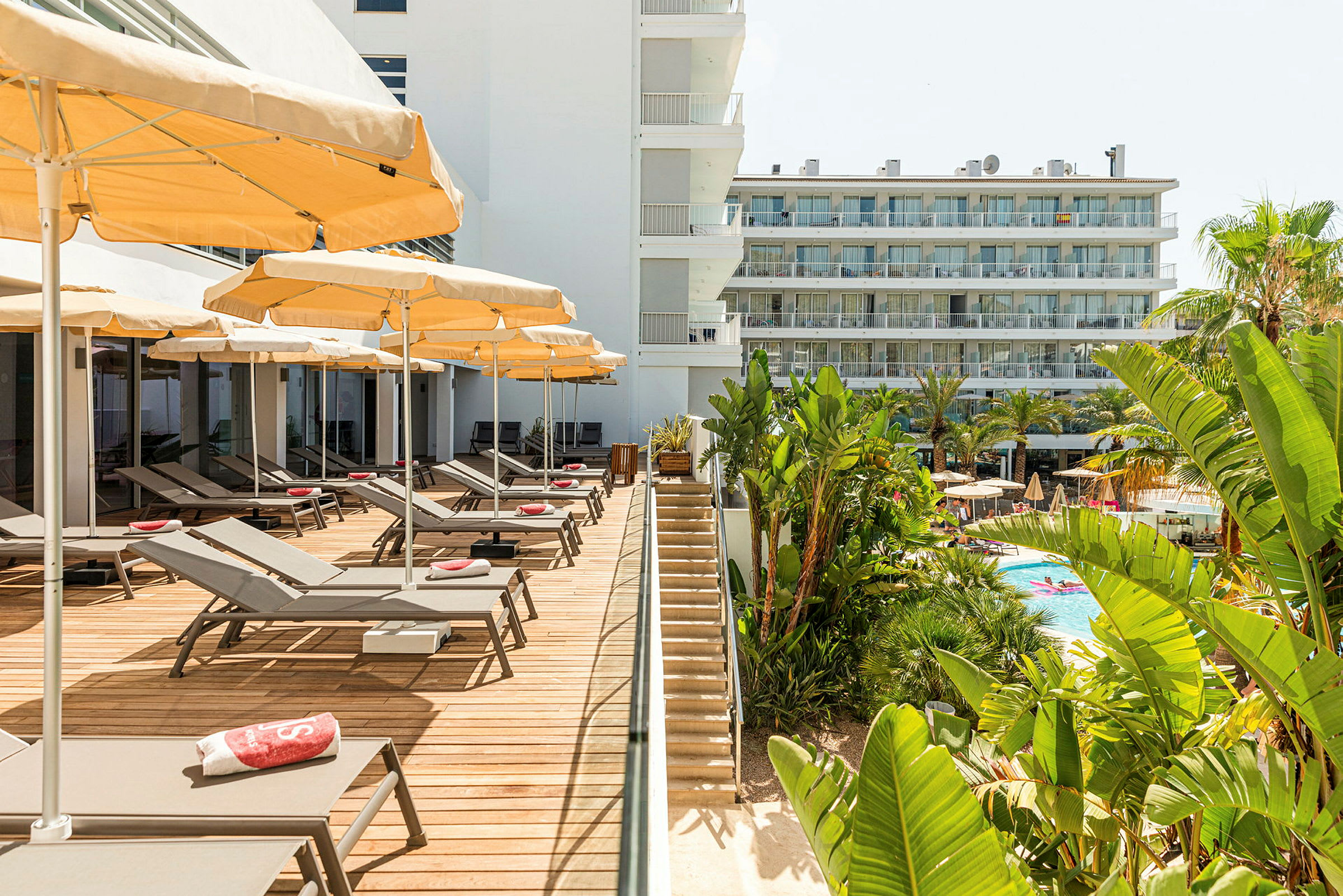 Et hotelområde med liggestole og parasoller på en træbeklædt terrasse ved siden af en trappe, der fører til poolområdet omgivet af frodig vegetation. (AI-genereret tekst)