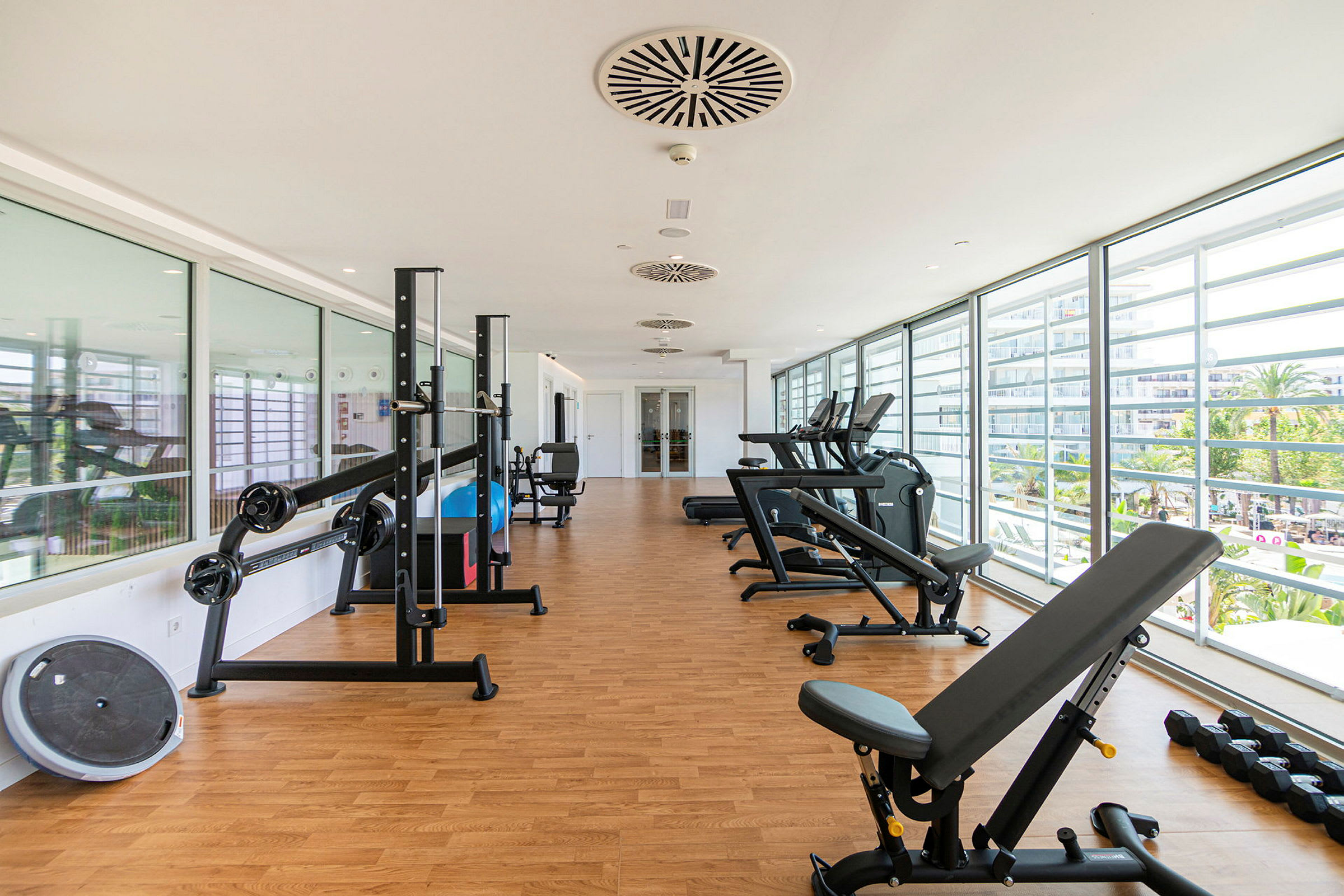 Et lyst fitnesscenter med træningsudstyr, store glasvinduer og udsigt til palmer. (AI-genereret tekst)