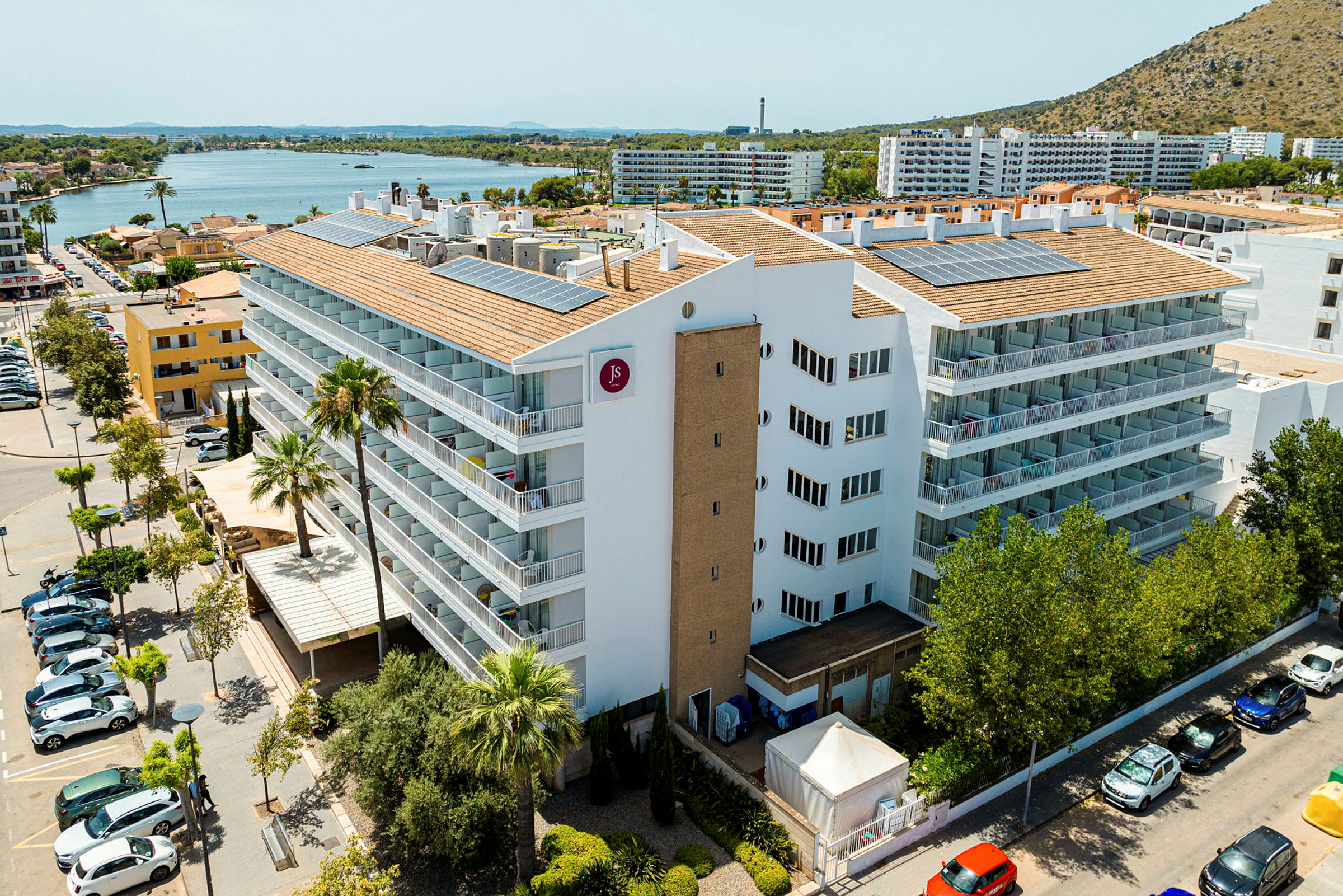 Hotelbygning ved en vej med parkerede biler, omgivet af træer og med udsigt over en sø. (AI-genereret tekst)