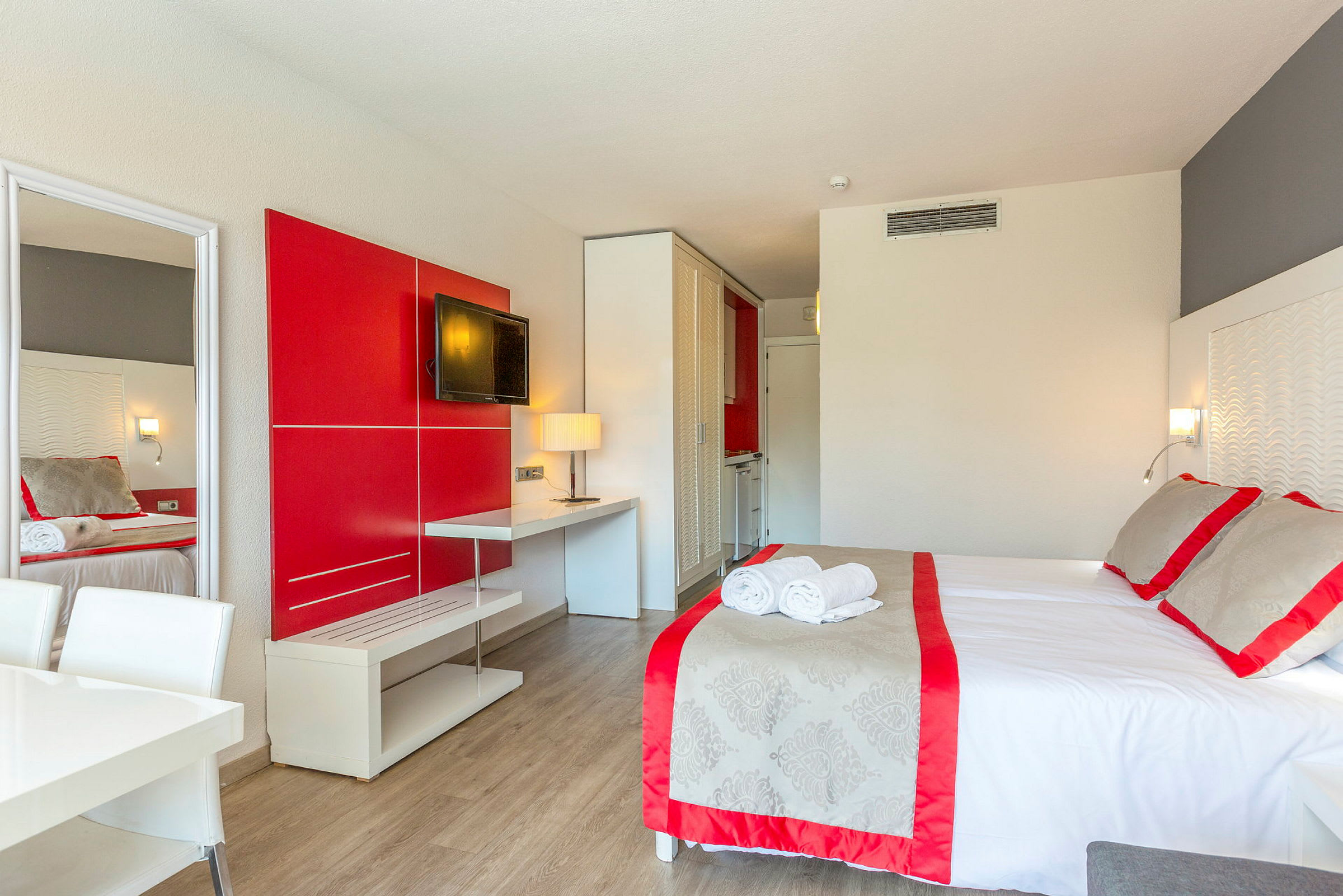 Et hotelværelse med en seng, røde og grå detaljer, et TV på væggen og et bord med en lampe. (AI-genereret tekst)