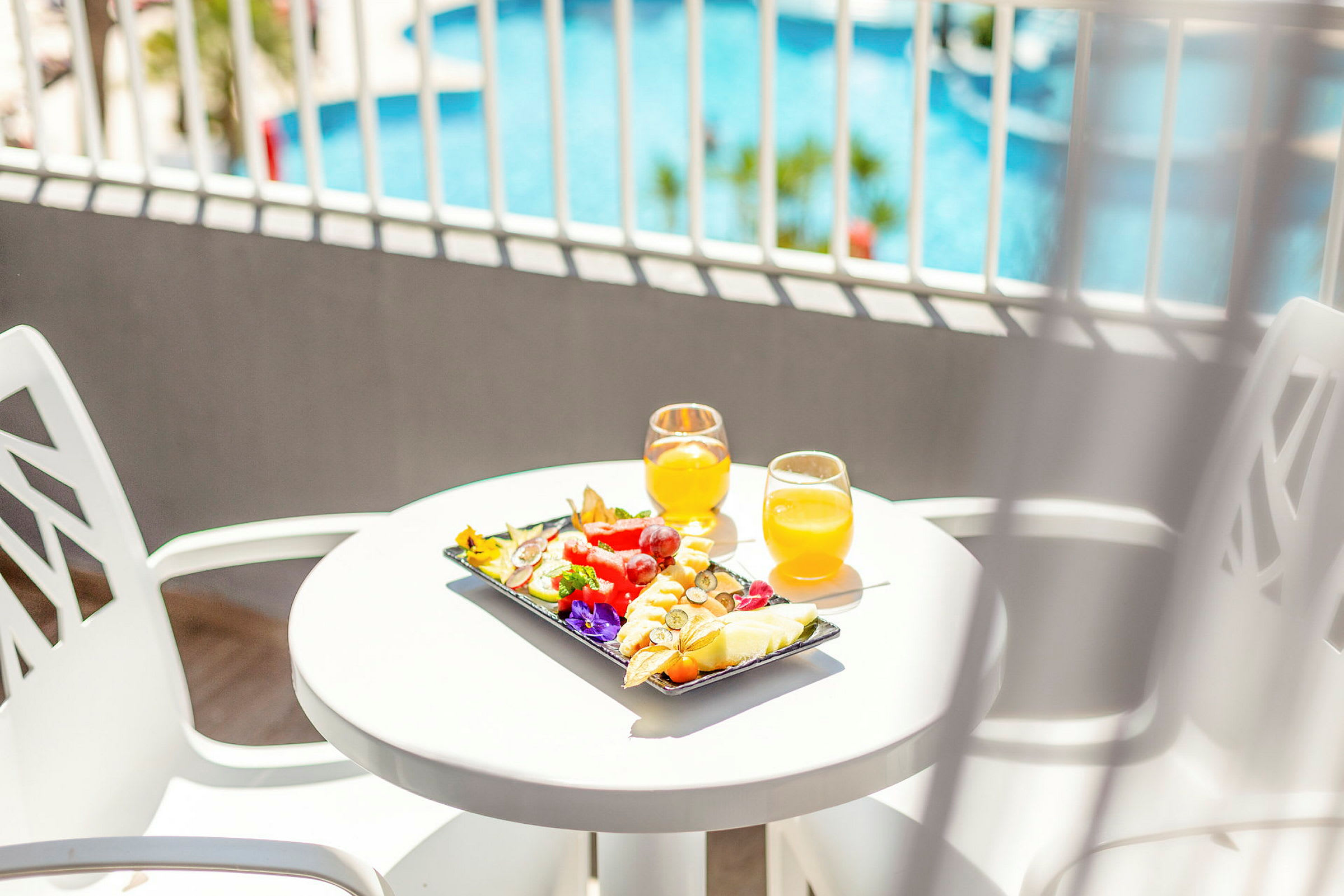 Et hvidt bord med frisk frugt og to glas appelsinjuice, med en pool i baggrunden. (AI-genereret tekst)