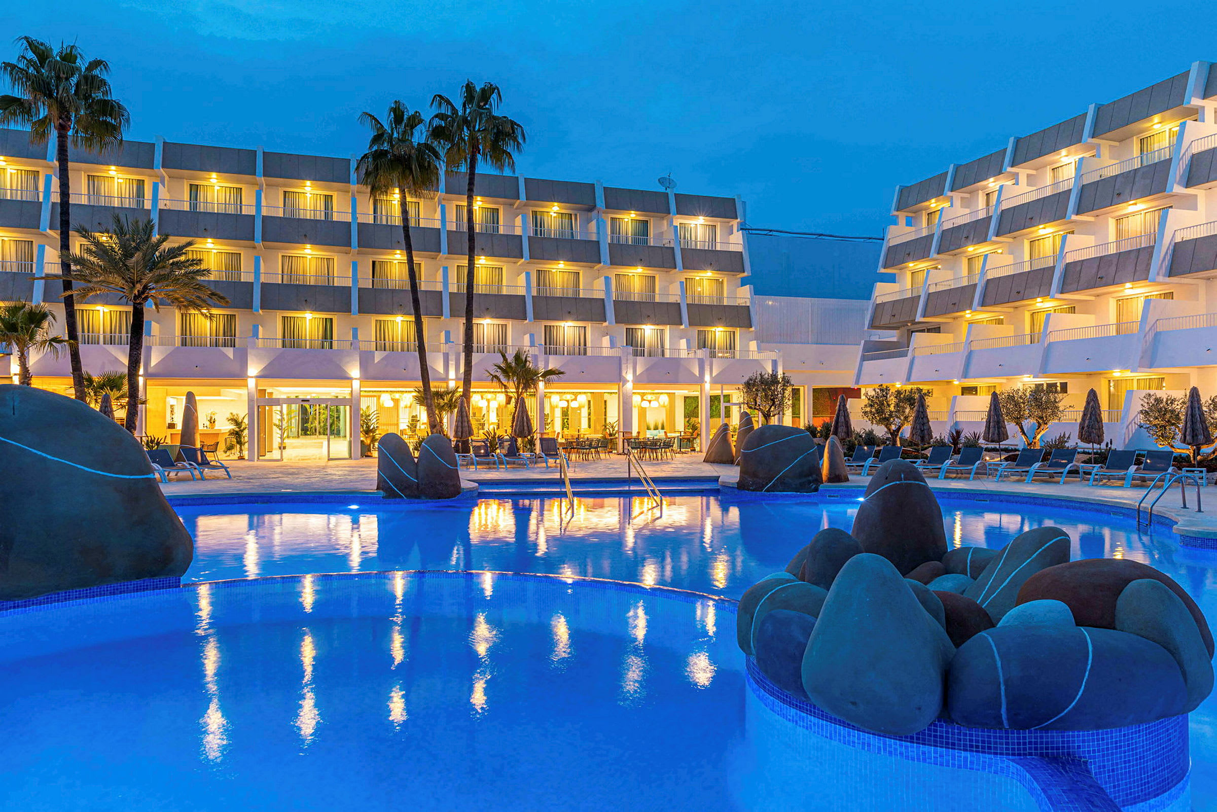 Hotellets facade i skumringen med oplyst poolområde og palmer. (AI-genereret tekst)