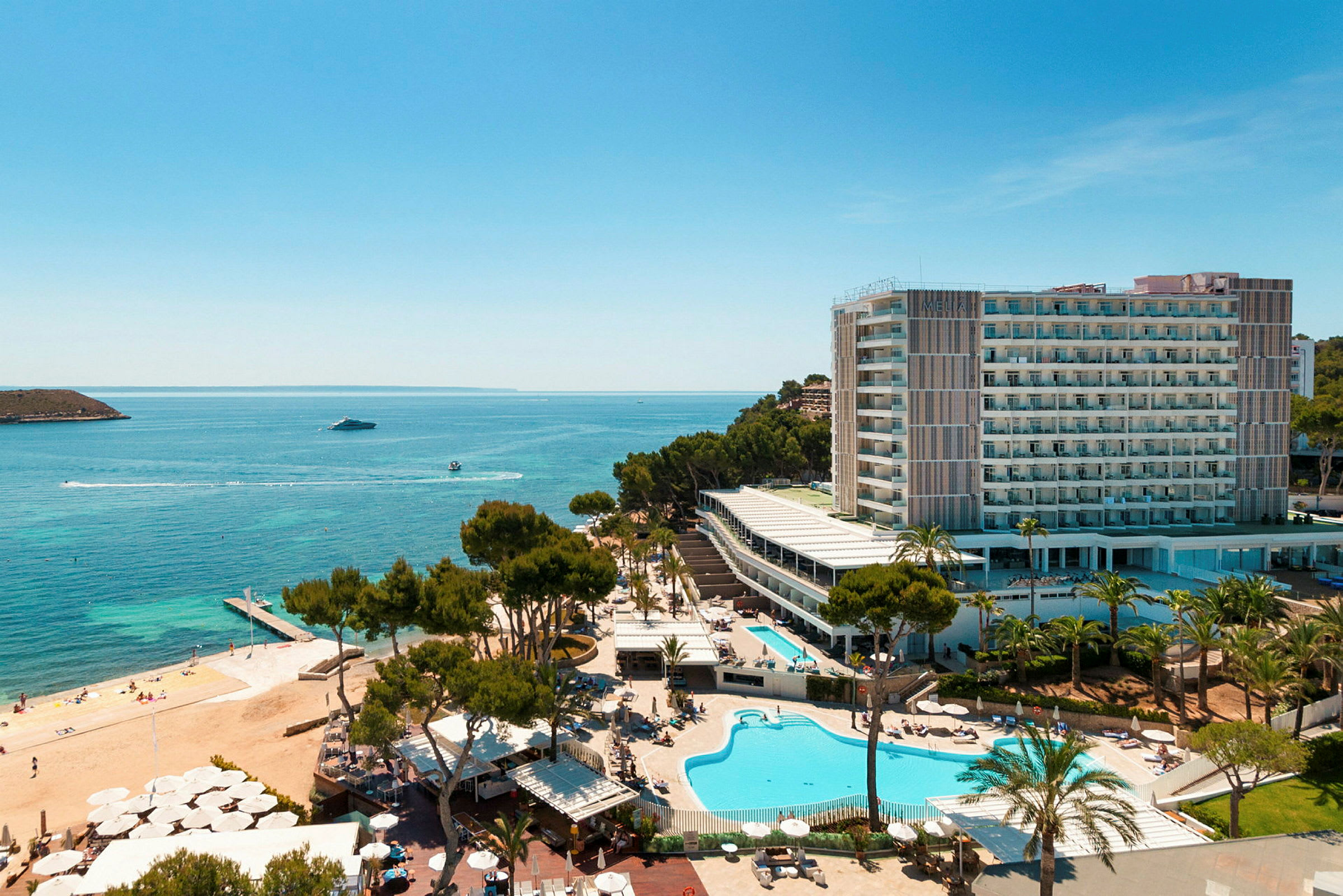 Luftfoto af et hotelkompleks ved havet med swimmingpools, grønne områder og en strand. (AI-genereret tekst)