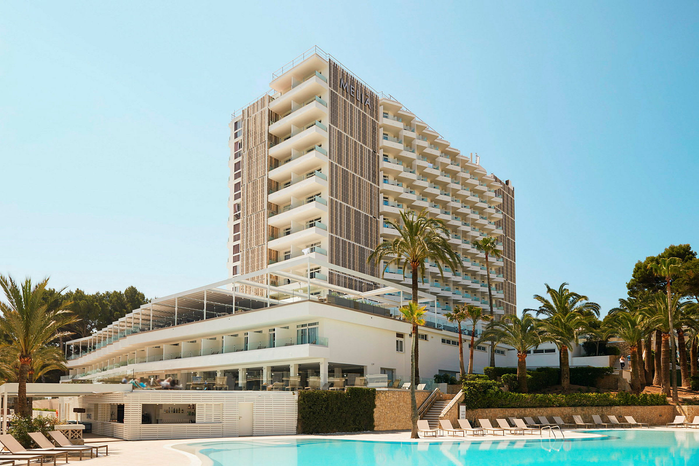 Et højhus hotel med moderne arkitektur beliggende ved siden af en udendørs pool omgivet af palmer. (AI-genereret tekst)