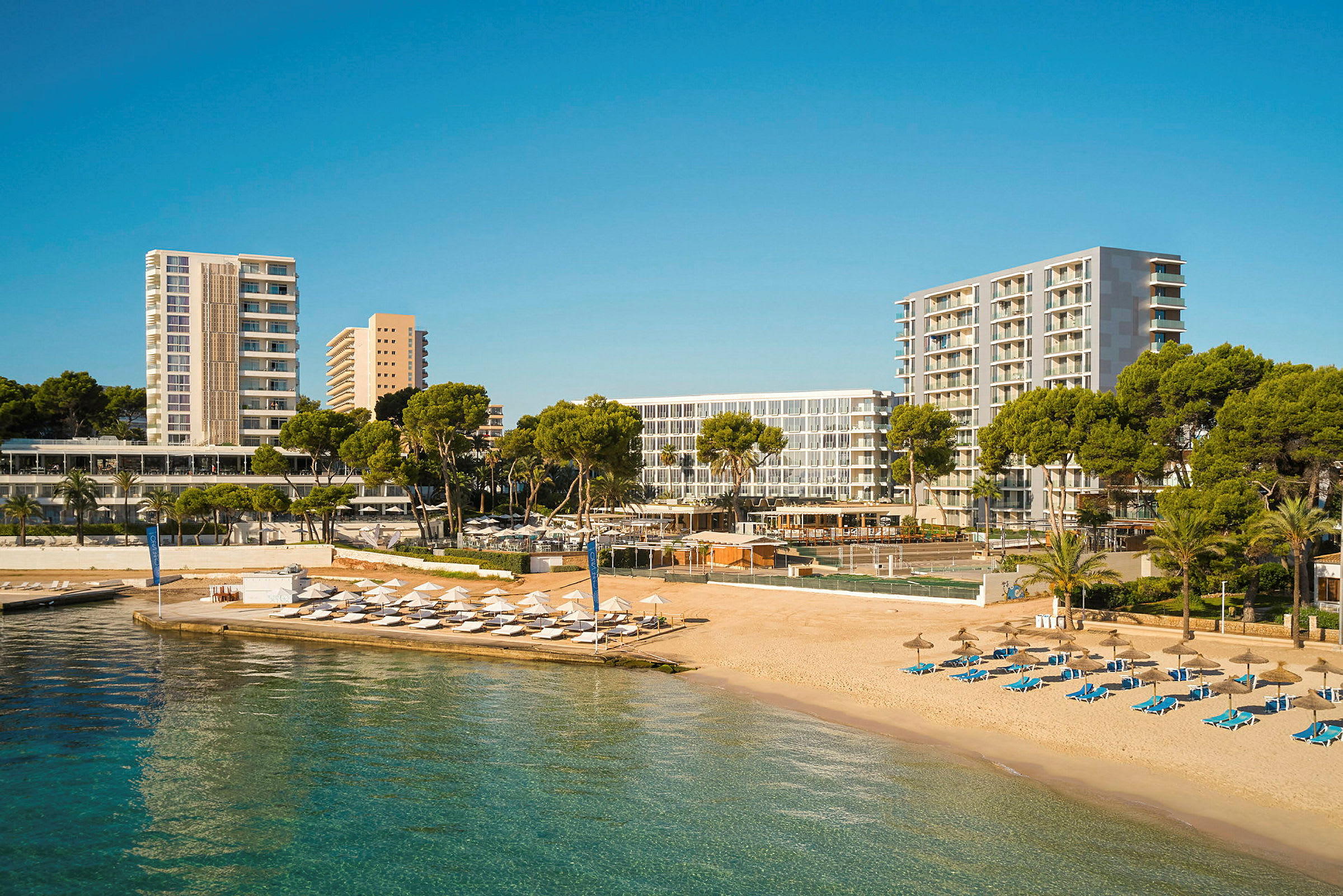 En strand med solsenge og parasoller ved siden af hotelbygninger og grønne områder. (AI-genereret tekst)