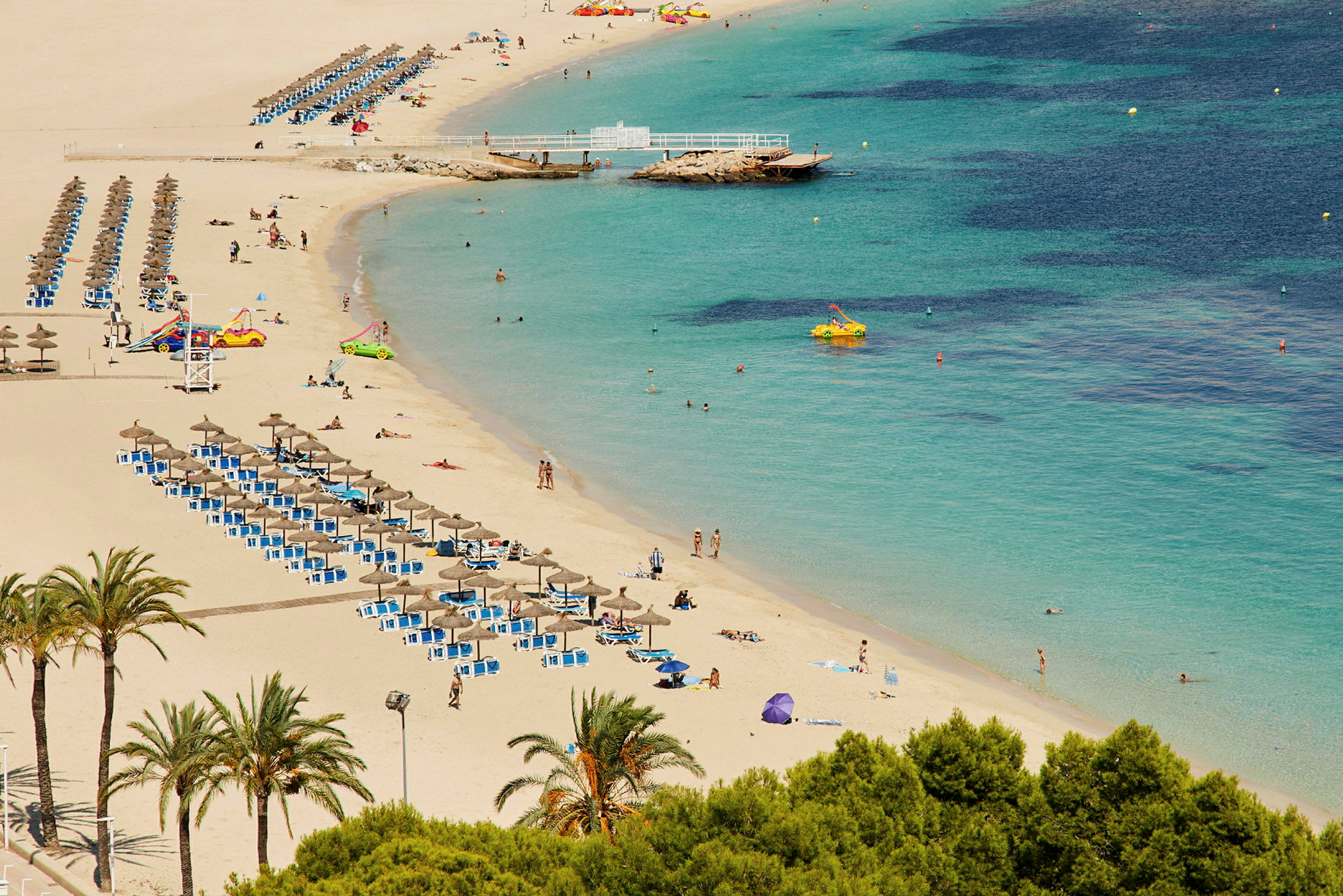 Strand med turkis vand, liggestole og parasoller på række samt palmer i forgrunden. (AI-genereret tekst)