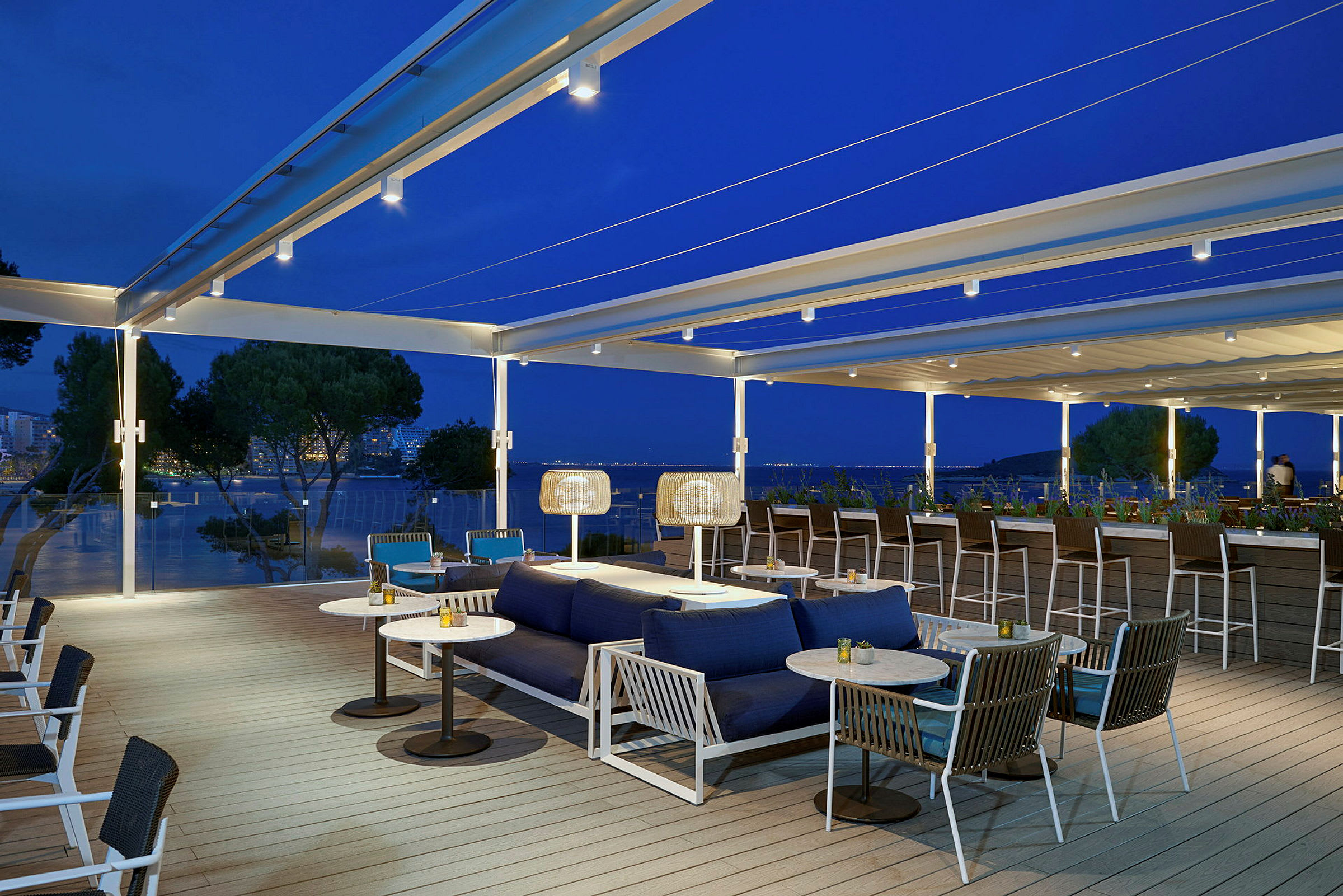 Billede af en udendørs terrasse med siddepladser og sofaer, placeret med en smuk udsigt over havet i skumringen. (AI-genereret tekst)