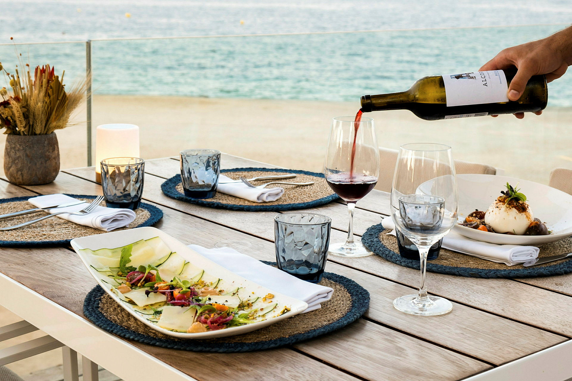 Et dækket bord med tallerkener, glas og flasker. Vin bliver skænket op i et glas med havet i baggrunden. (AI-genereret tekst)