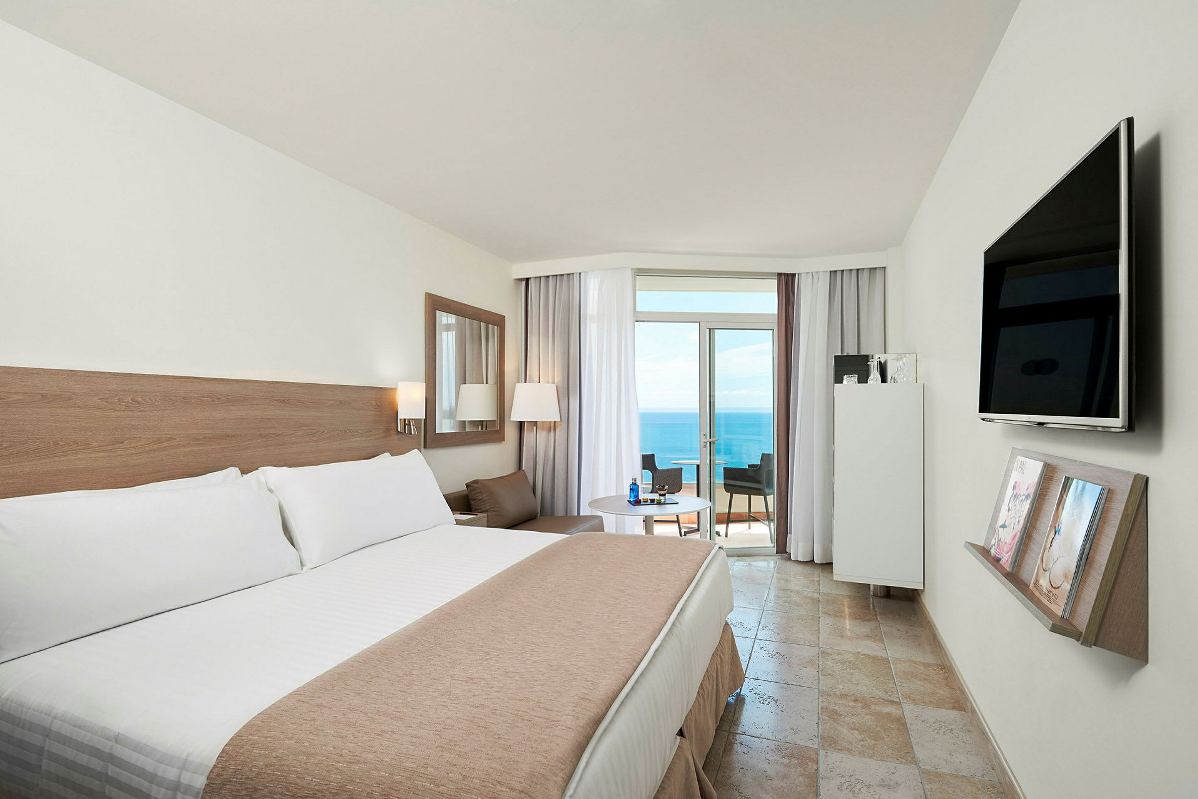 Hotelværelse med en stor seng, et bord med stole og en balkon med udsigt over havet. (AI-genereret tekst)