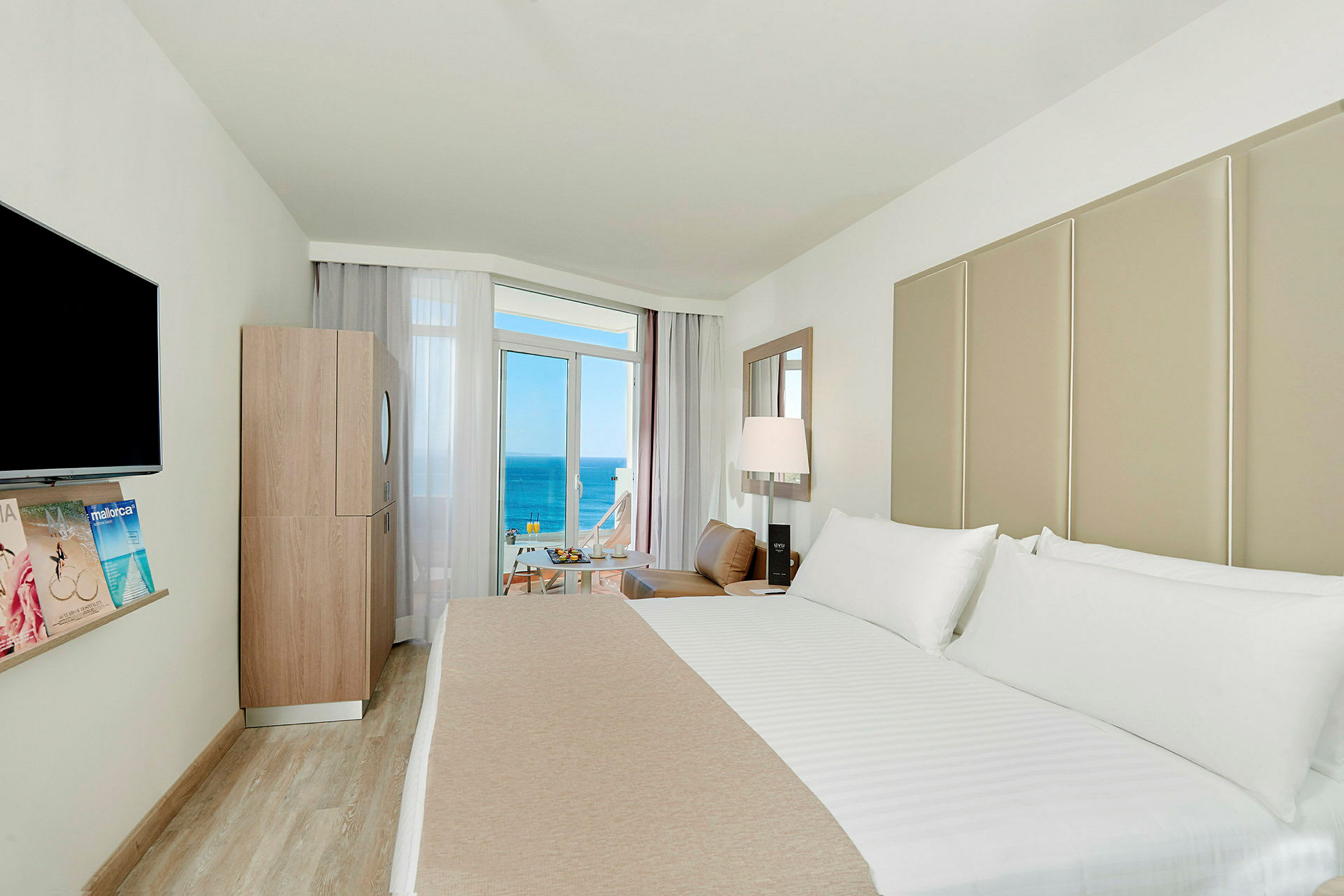 Et hotelværelse med en stor seng, et TV på væggen, en moderne indretning og en balkon med udsigt over havet. (AI-genereret tekst)