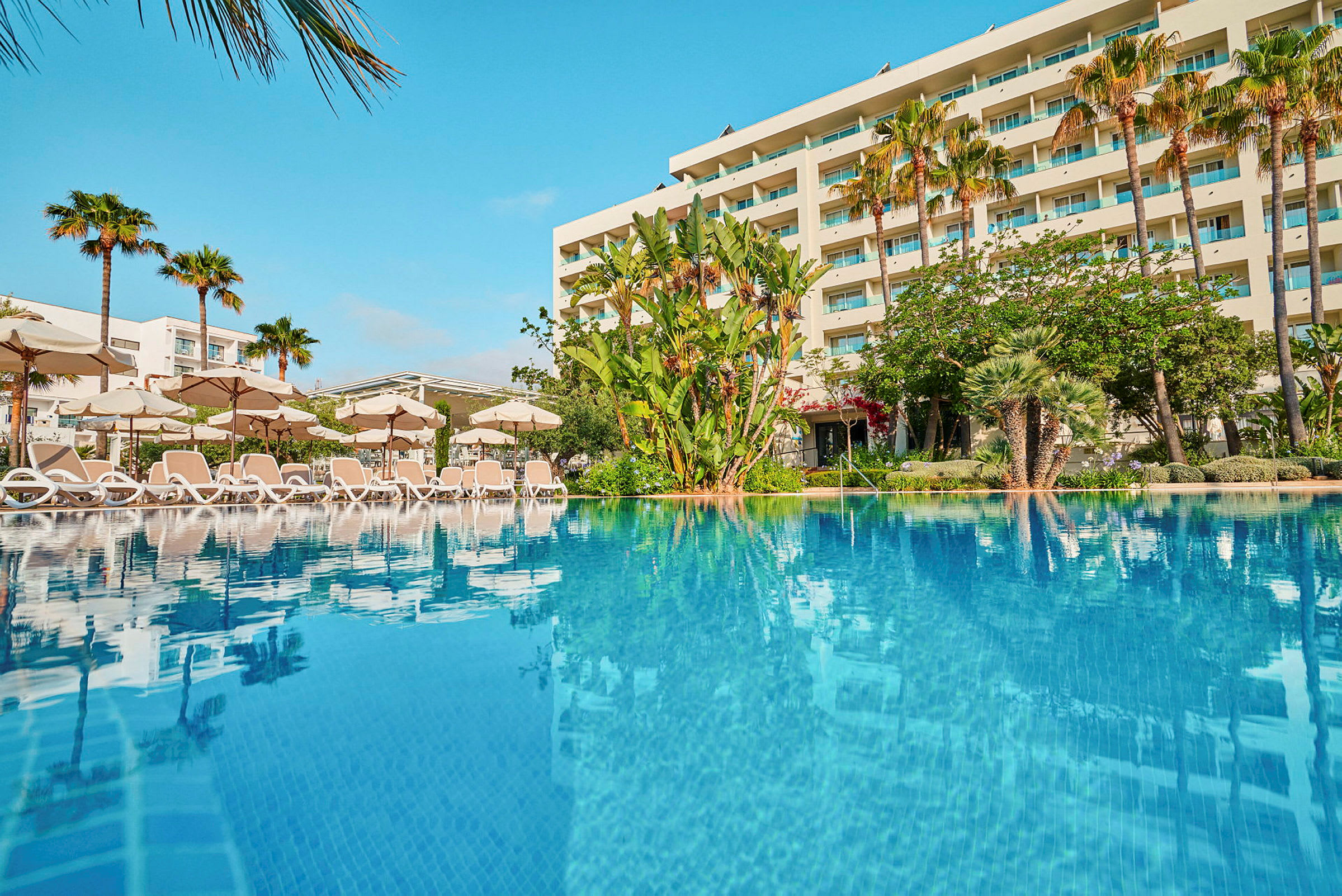 Hotel med pool og liggestole foran en have og palmer. (AI-genereret tekst)