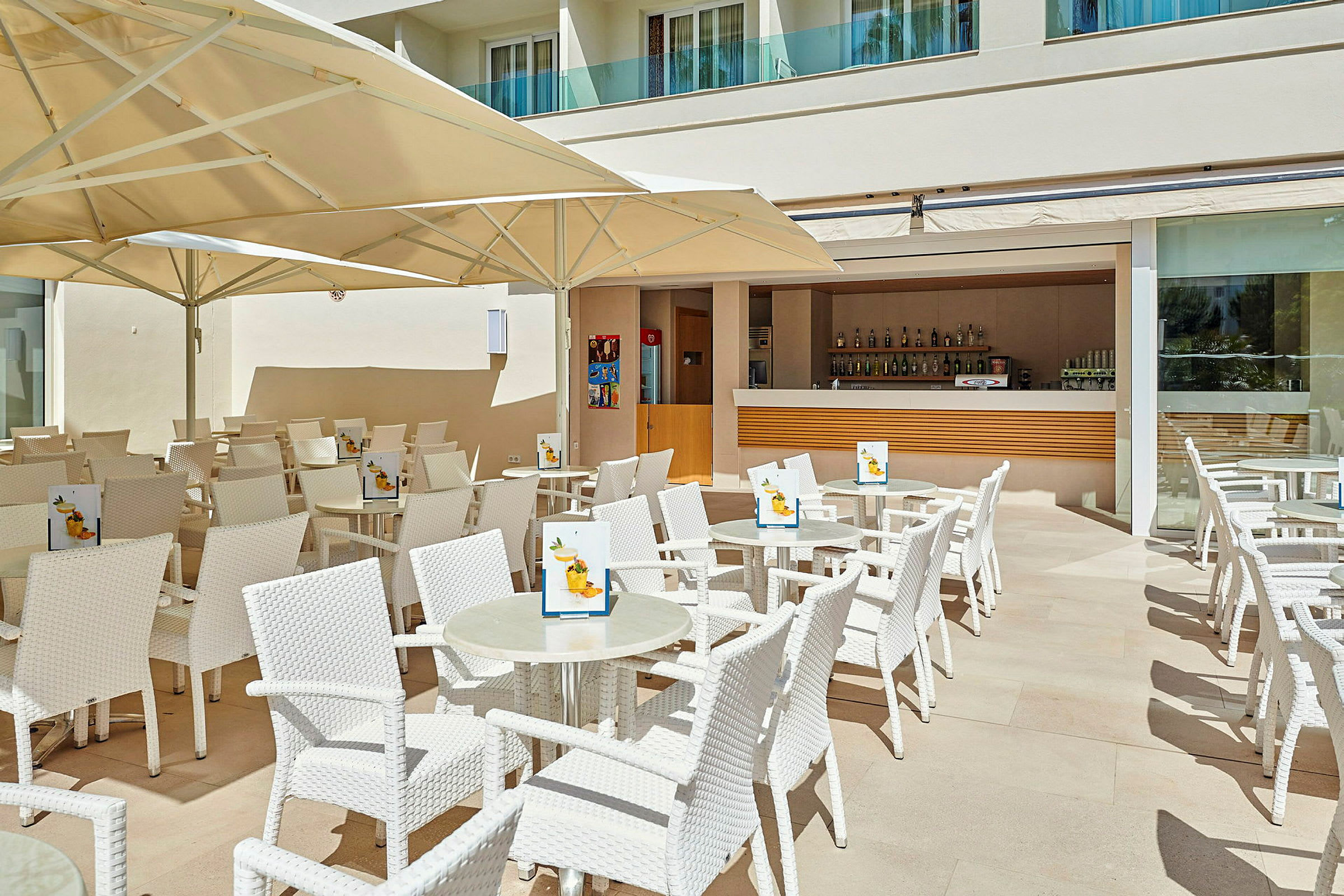Terrasse med hvide stole og borde, under store parasoller, foran en åben bar. (AI-genereret tekst)