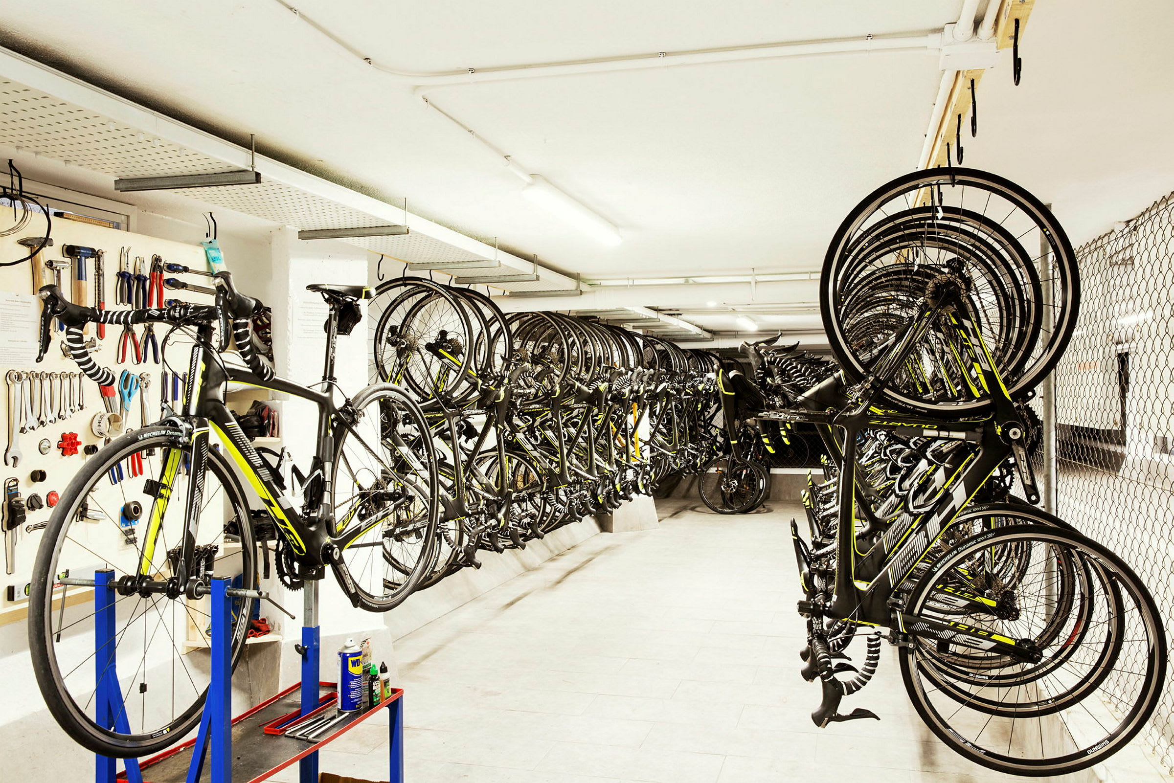 Cykelrum med værktøjsplade, cykel på monteringsstander og mange racercykler hængende. (AI-genereret tekst)