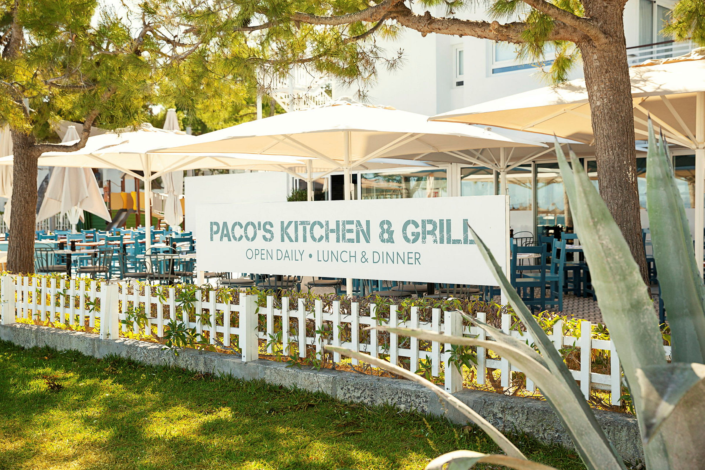 Skilt med teksten 'Paco's Kitchen & Grill' foran en restaurant med udendørs servering. (AI-genereret tekst)