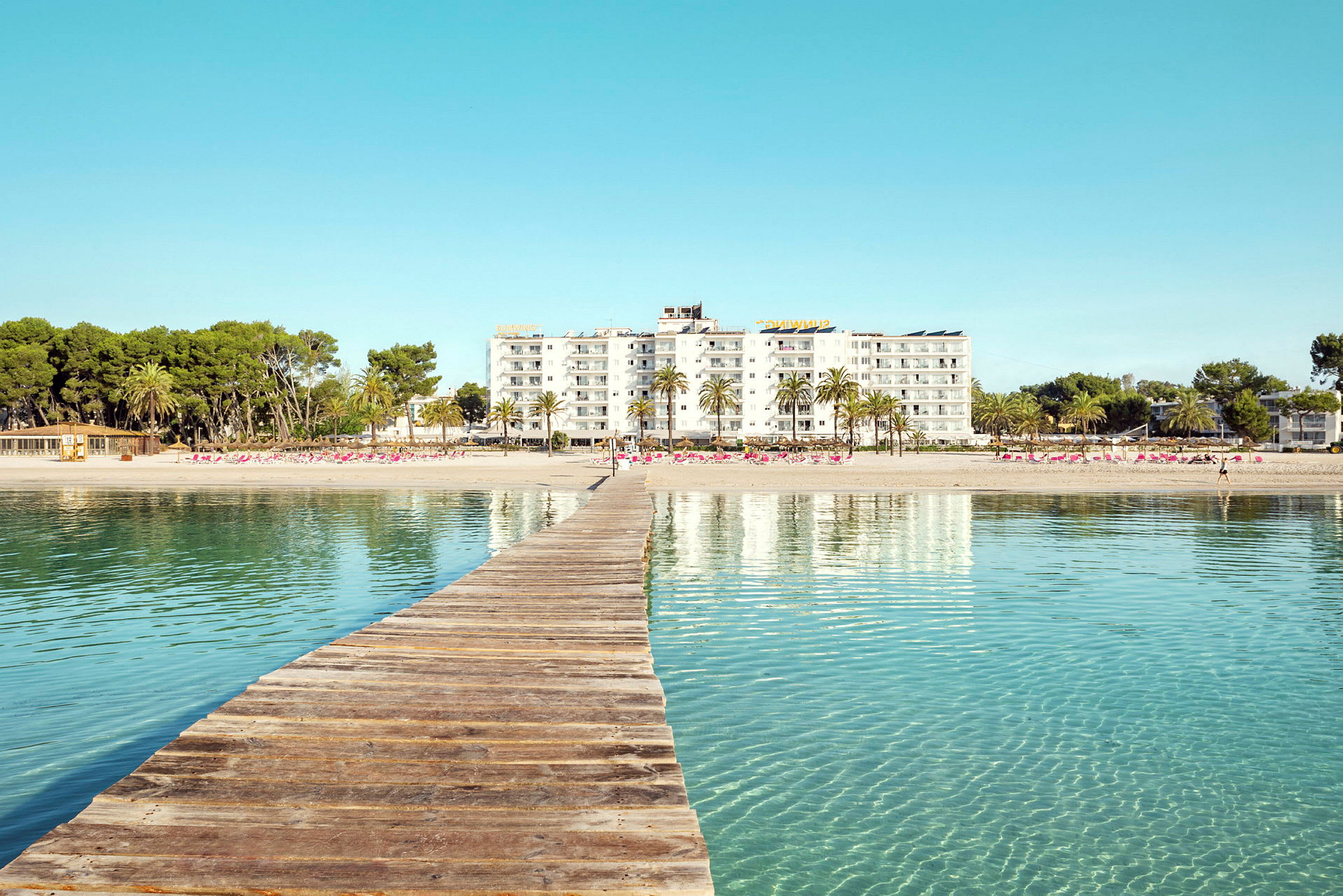 Træbro, der fører mod en sandstrand med hotelbygninger og palmer i baggrunden. (AI-genereret tekst)