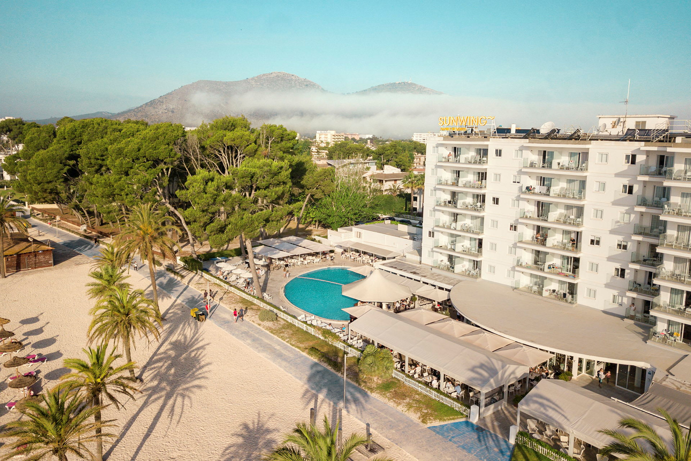 Hotelområde med pool, omgivet af grønne områder og med stranden i forgrunden. (AI-genereret tekst)