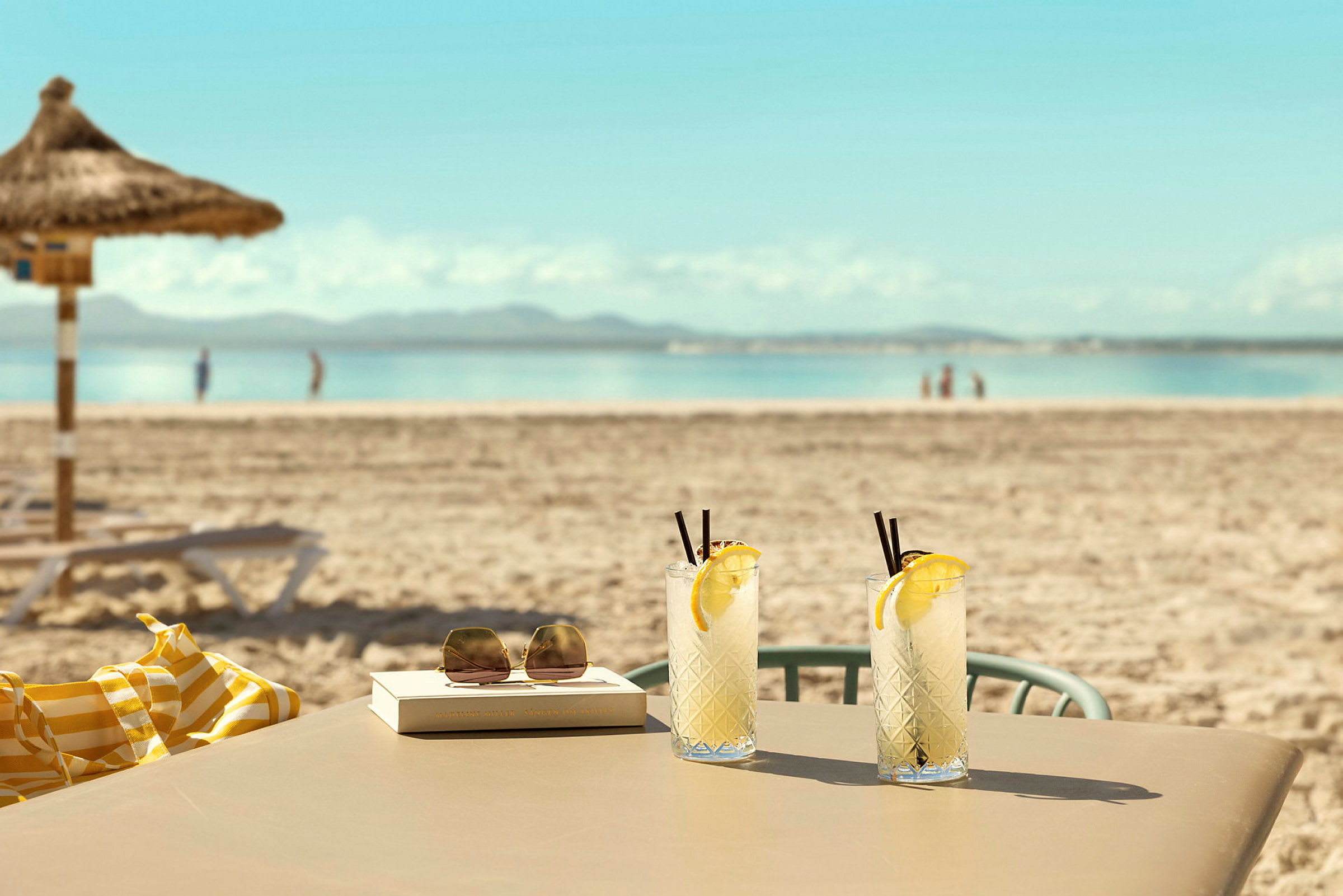 To glas med limonade står på et bord ved en sandstrand med havet i baggrunden. (AI-genereret tekst)
