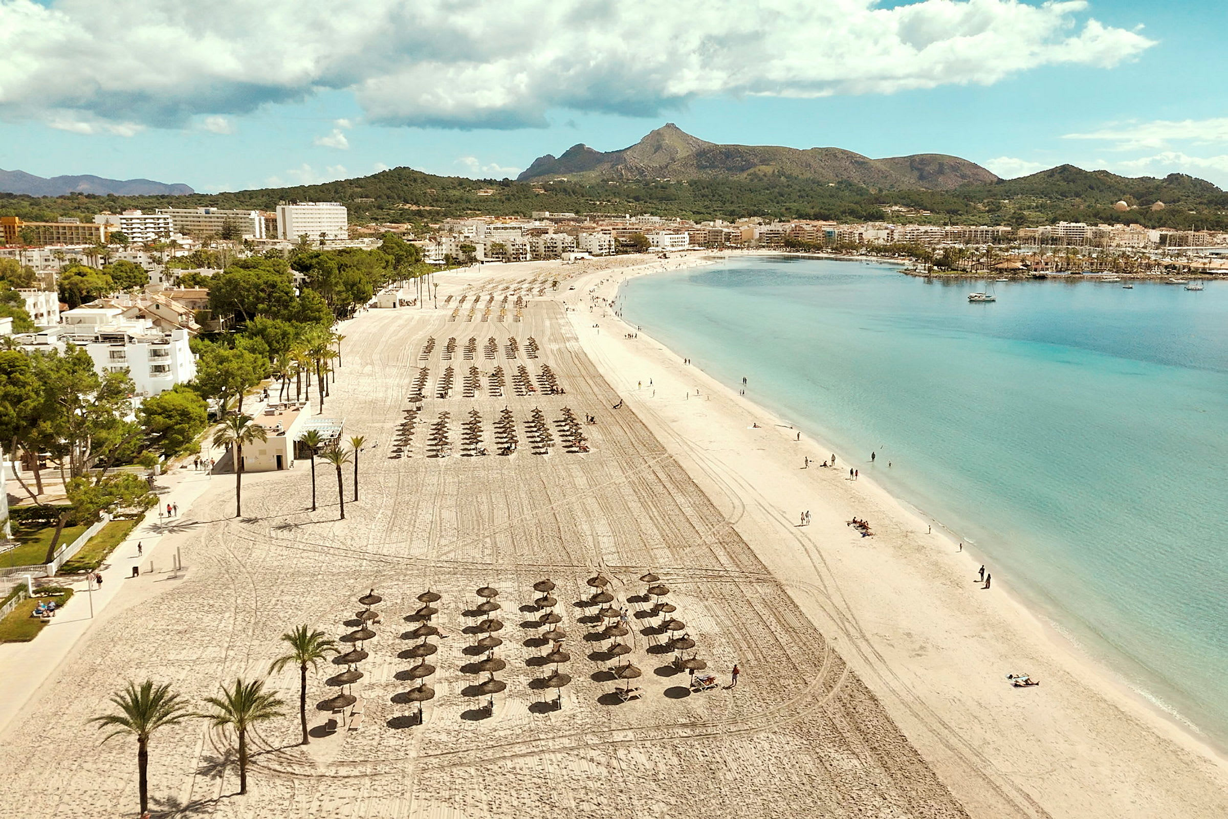 En langstrakt sandstrand med parasoller og klart blåt hav. I baggrunden ses bygninger og grønne bjerge. (AI-genereret tekst)