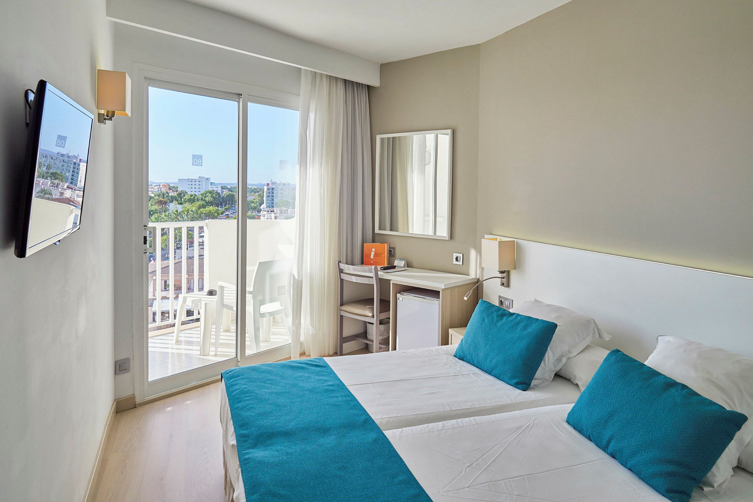Et lyst hotelværelse med to senge, blå puder og sengetæpper samt en balkon med udsigt. (AI-genereret tekst)