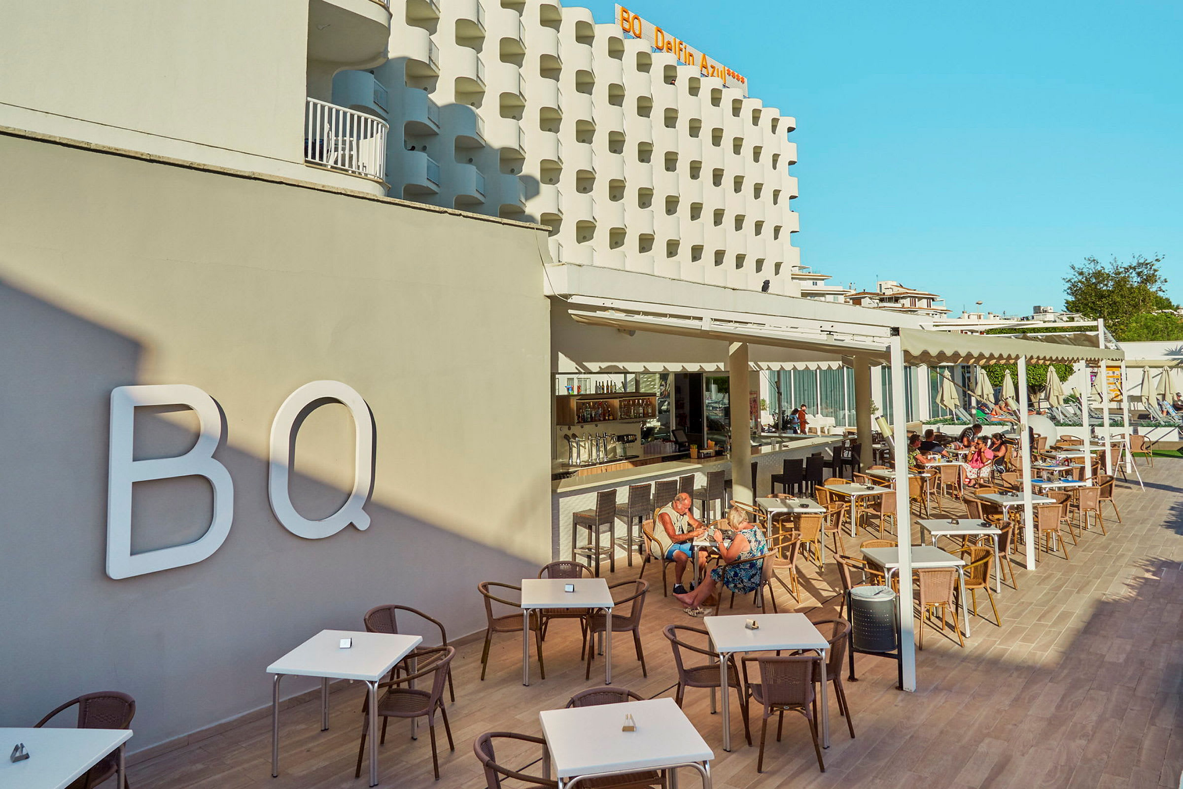 Hotellets facade og udendørs serveringsområde med borde og stole, mennesker der sidder og spiser. (AI-genereret tekst)