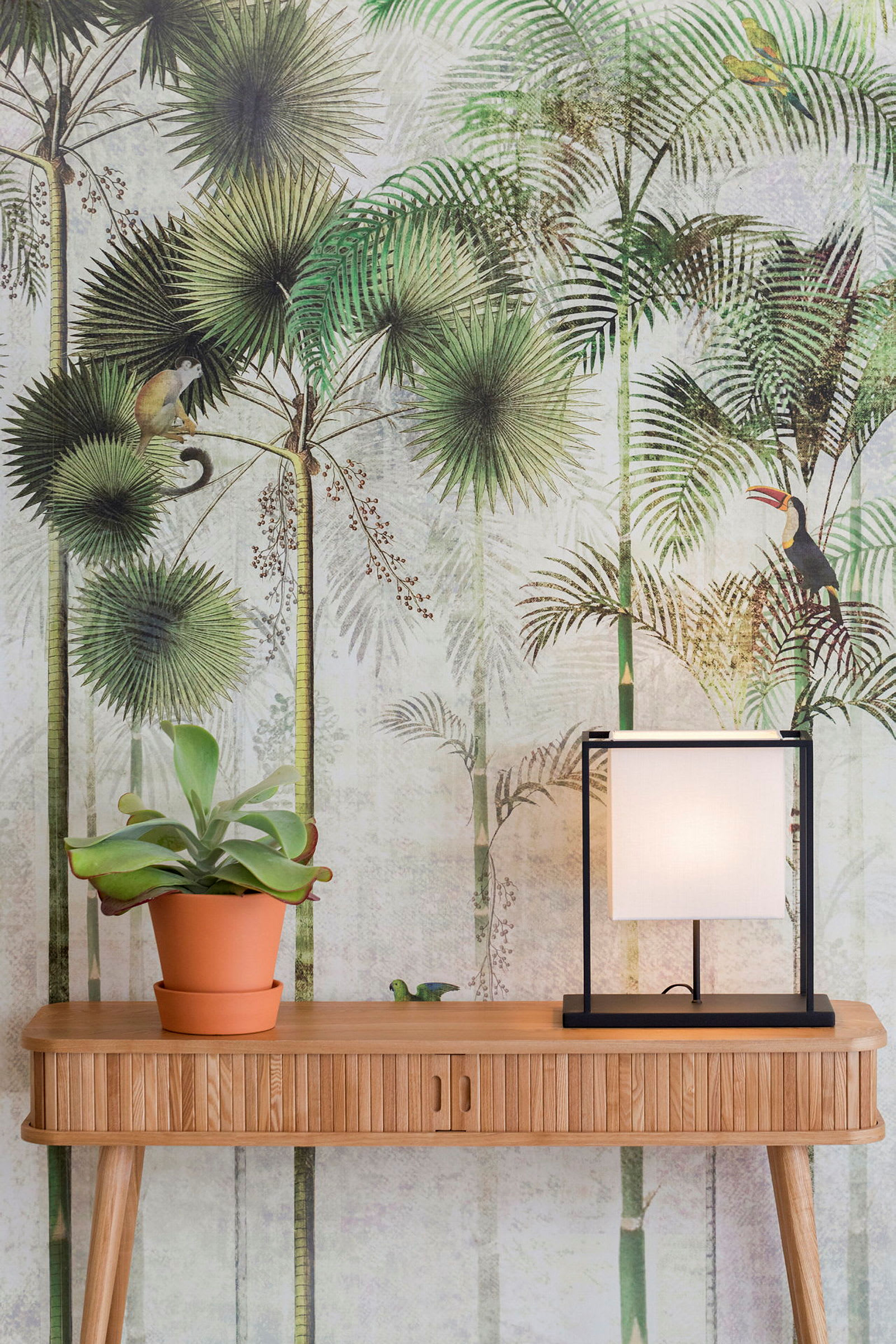 En træbord med en potteplante og en firkantet lampe; i baggrunden er der en tropisk tapet med dyre- og plantemotiver. (AI-genereret tekst)